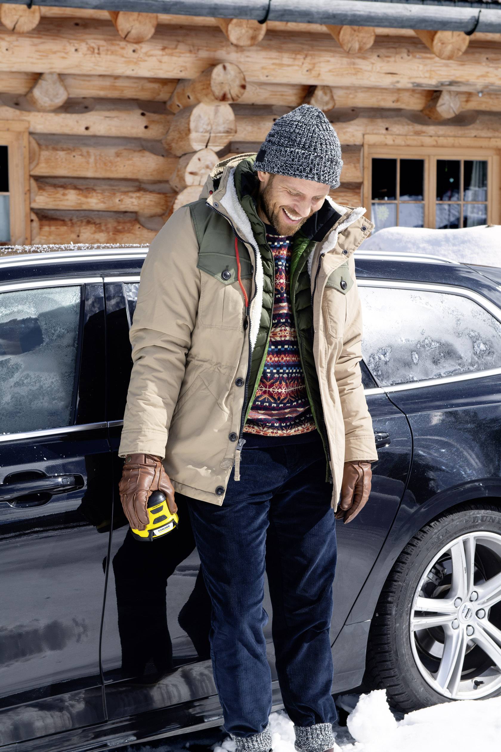 Ein Mann lächelt neben einem Auto im Schnee, warm gekleidet mit Mütze und Jacke, hält ein gelbes Gerät in der Hand. Eine Holzhütte im Hintergrund.