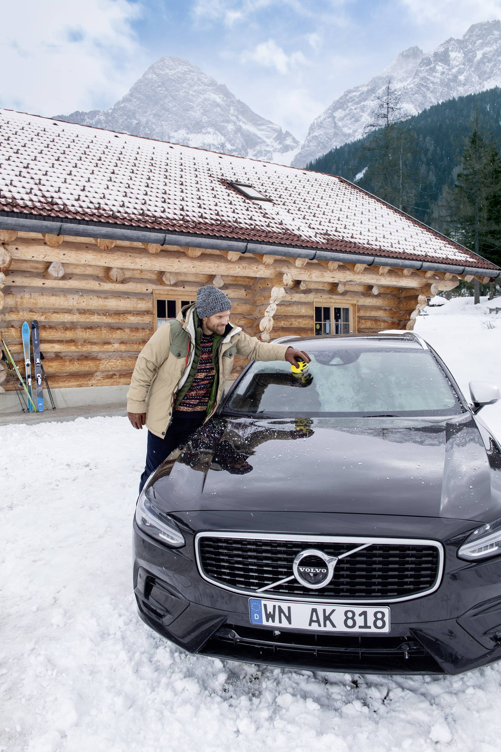 Ein Mann in Winterkleidung wischt Schnee von einem schwarzen Auto vor einer Holzhütte und einer schneebedeckten Berglandschaft.