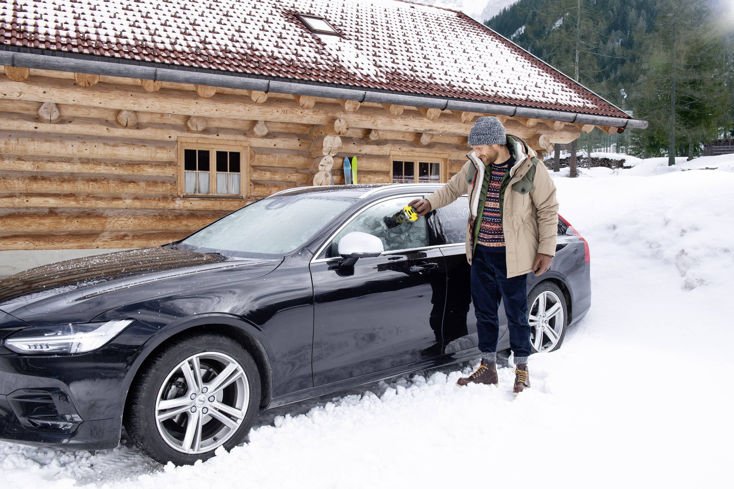 Ein Mann im Wintermantel steht vor einem verschneiten Auto und entfernt Schnee vom Fenster. Eine Holzhütte ist im Hintergrund zu sehen.