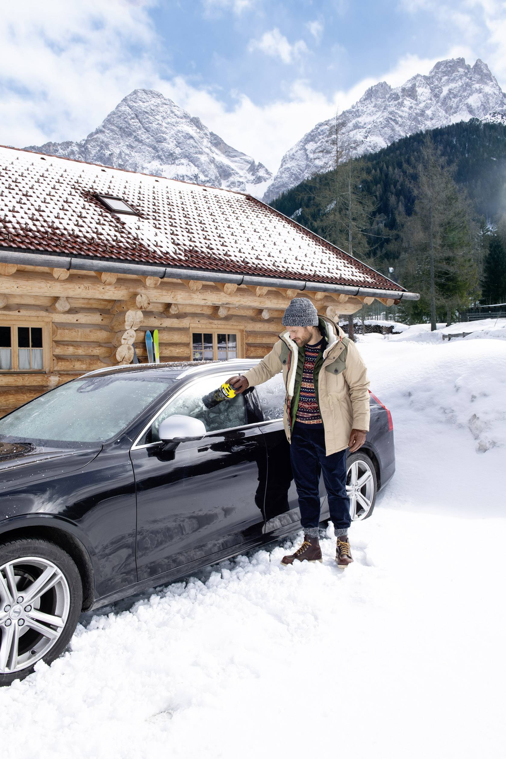 Eine Person mit Winterkleidung entfernt Schnee von einem schwarzen Auto vor einer Holzhütte in einer verschneiten Berglandschaft.