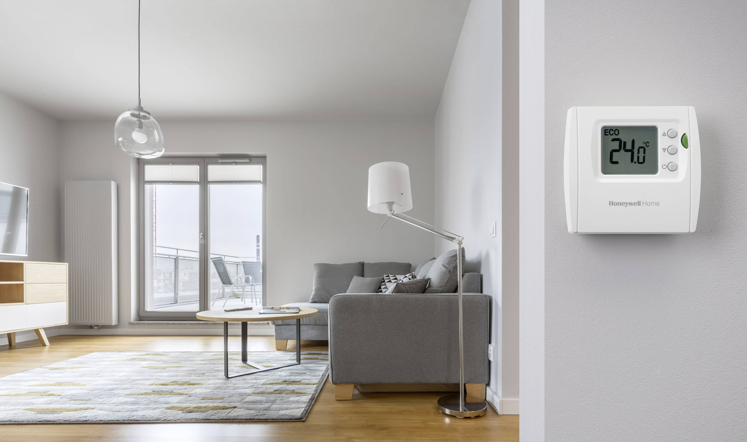 Wohnzimmer mit grauem Sofa, Lampe und Teppich. An der Wand ein digitales Thermostat, zeigt '24°C', im Eco-Modus. Balkon im Hintergrund.