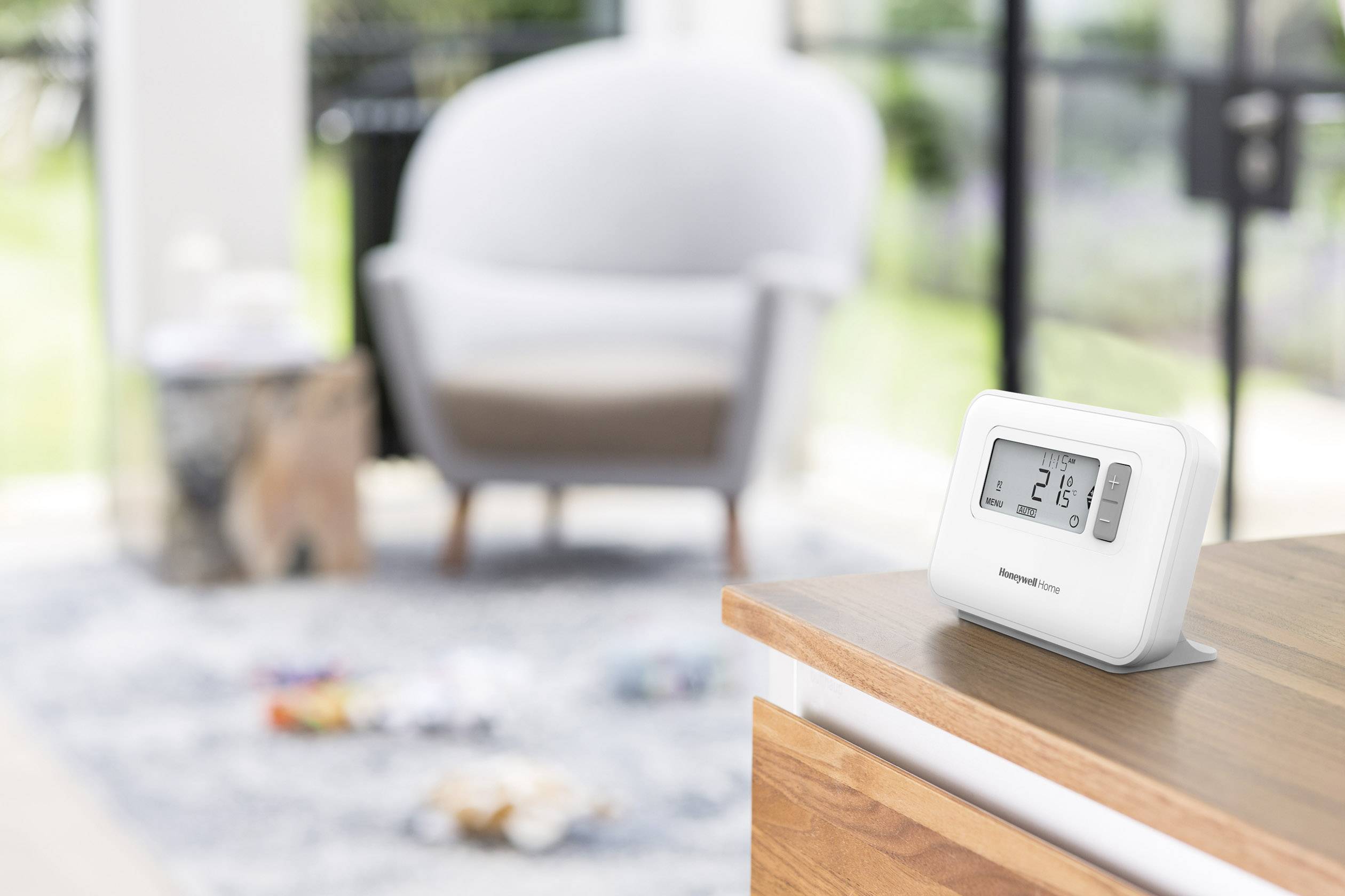 Ein digitales Raumthermostat zeigt 21 Grad Celsius an und steht auf einer Holzoberfläche. Im Hintergrund verschwommene Möbel.