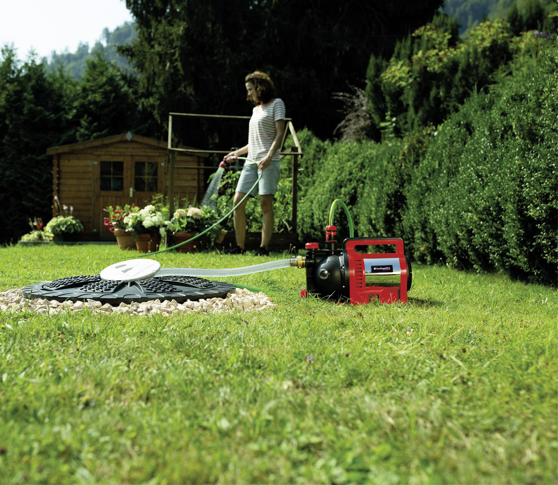 Eine Person bewässert den Garten mit einem roten Wasserdrucksystem. Im Hintergrund ist ein Holzschuppen und Pflanzen zu sehen.