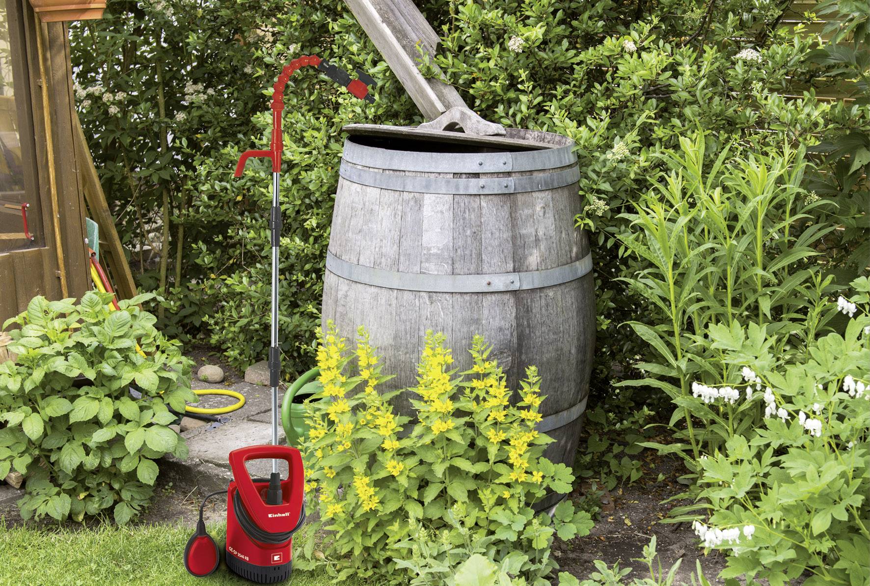 Ein alter Holzfass mit einem offenen Deckel steht in einem Garten, umgeben von grünen Pflanzen und einem roten Wasserpumpengerät daneben.