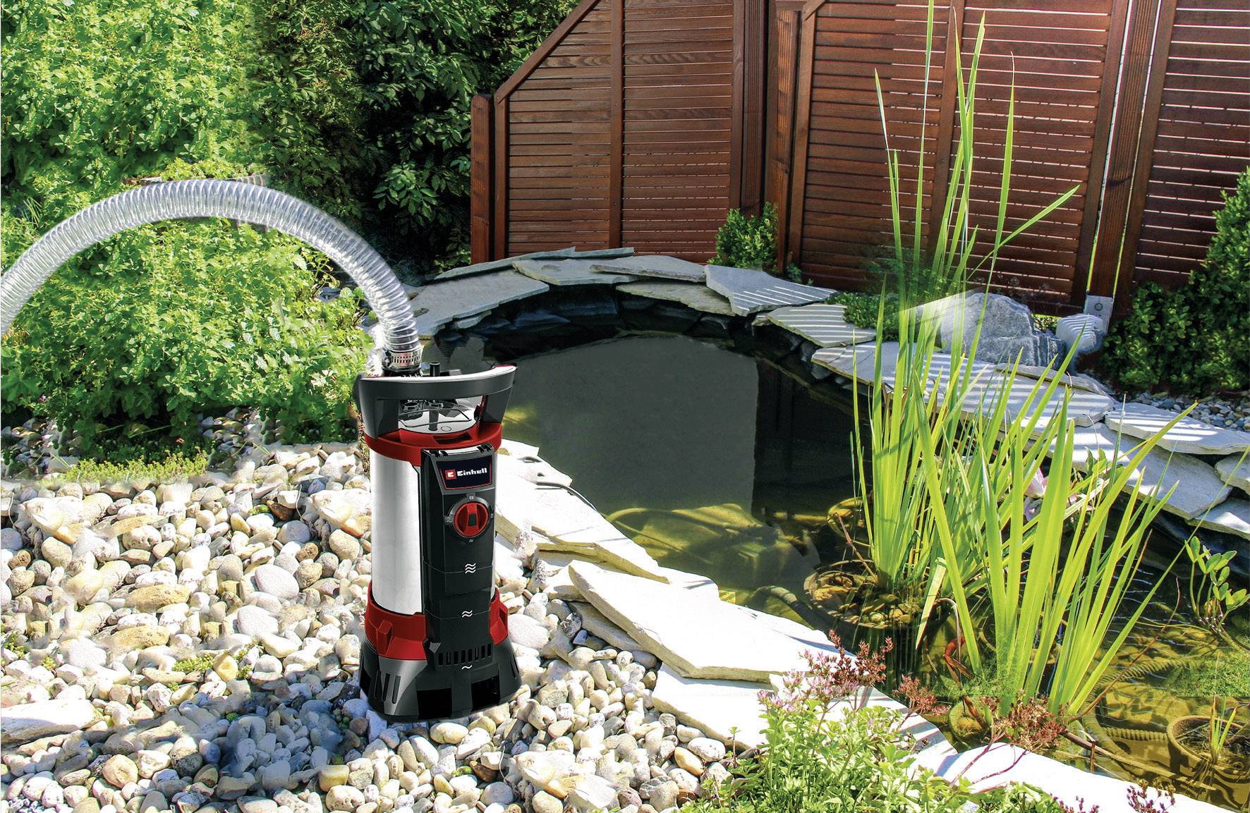 Ein Teichfilter mit einem Schlauch ist neben einem Steinrand-Teich mit Pflanzen und Holzumzäunung platziert, um Wasser sauber zu halten.