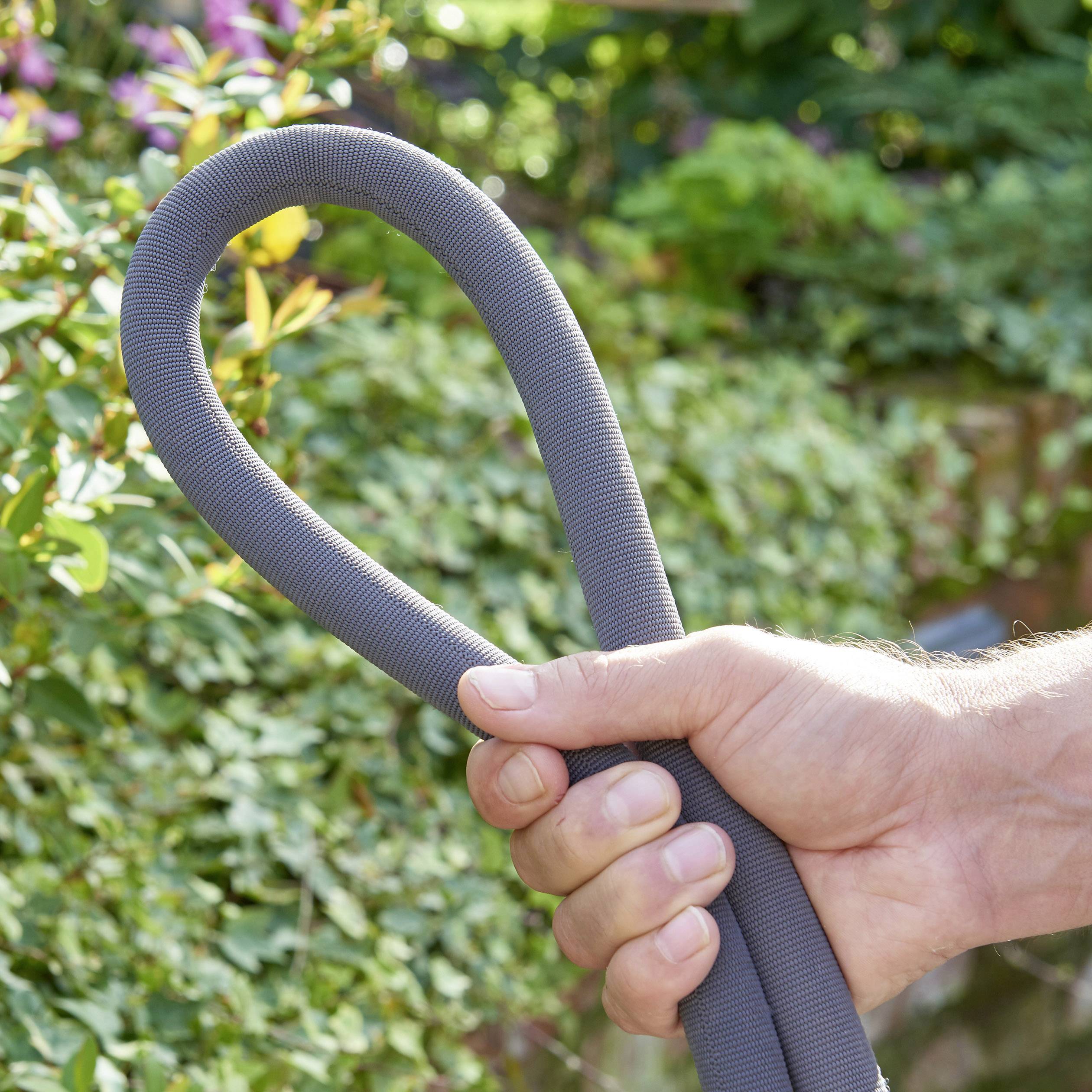 Hand hält einen grauen, flexiblen Gartenschlauch in einer grünen Gartenumgebung. Hintergrund zeigt Pflanzen in Unschärfe.