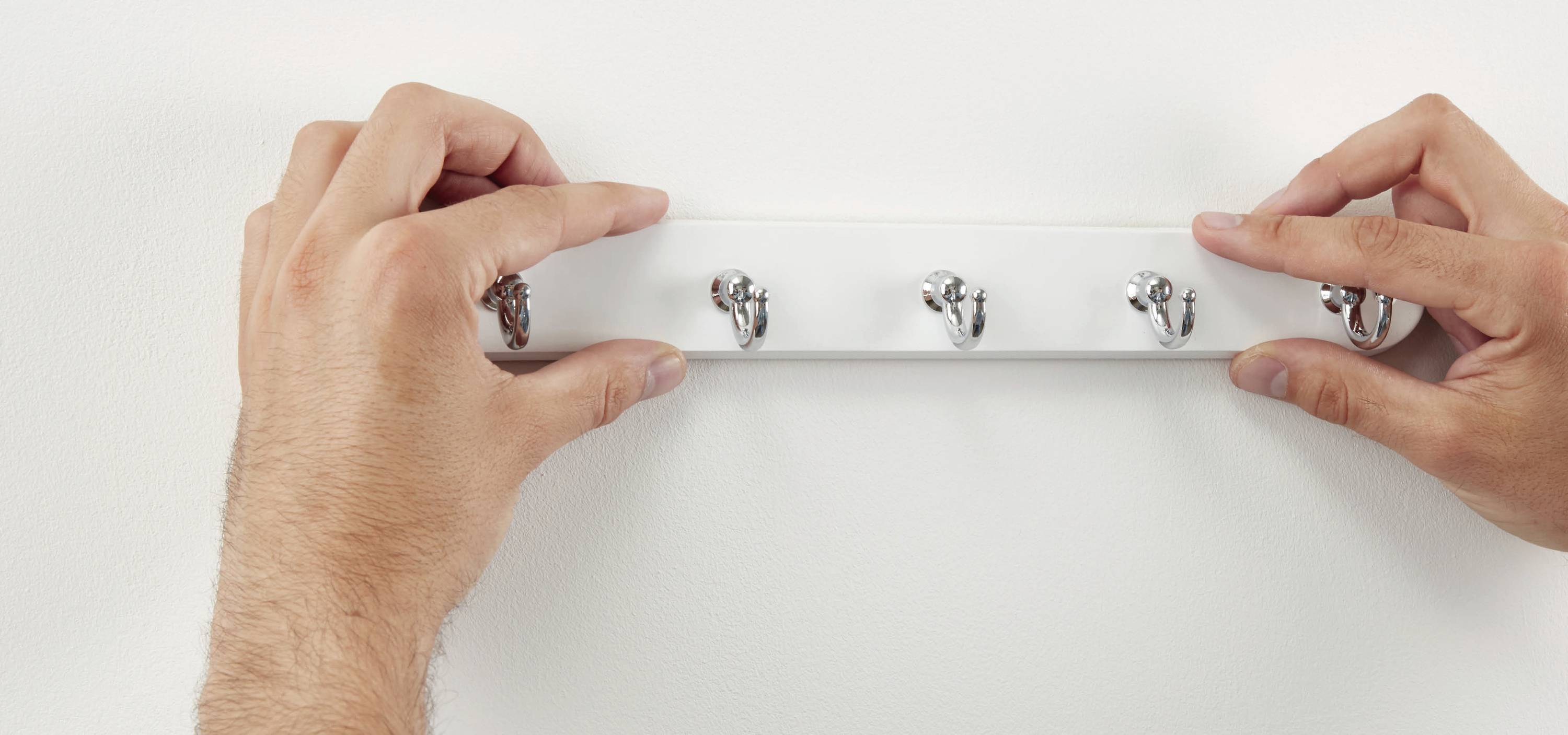 Hände montieren eine weiße Hakenleiste mit fünf Haken an einer Wand.