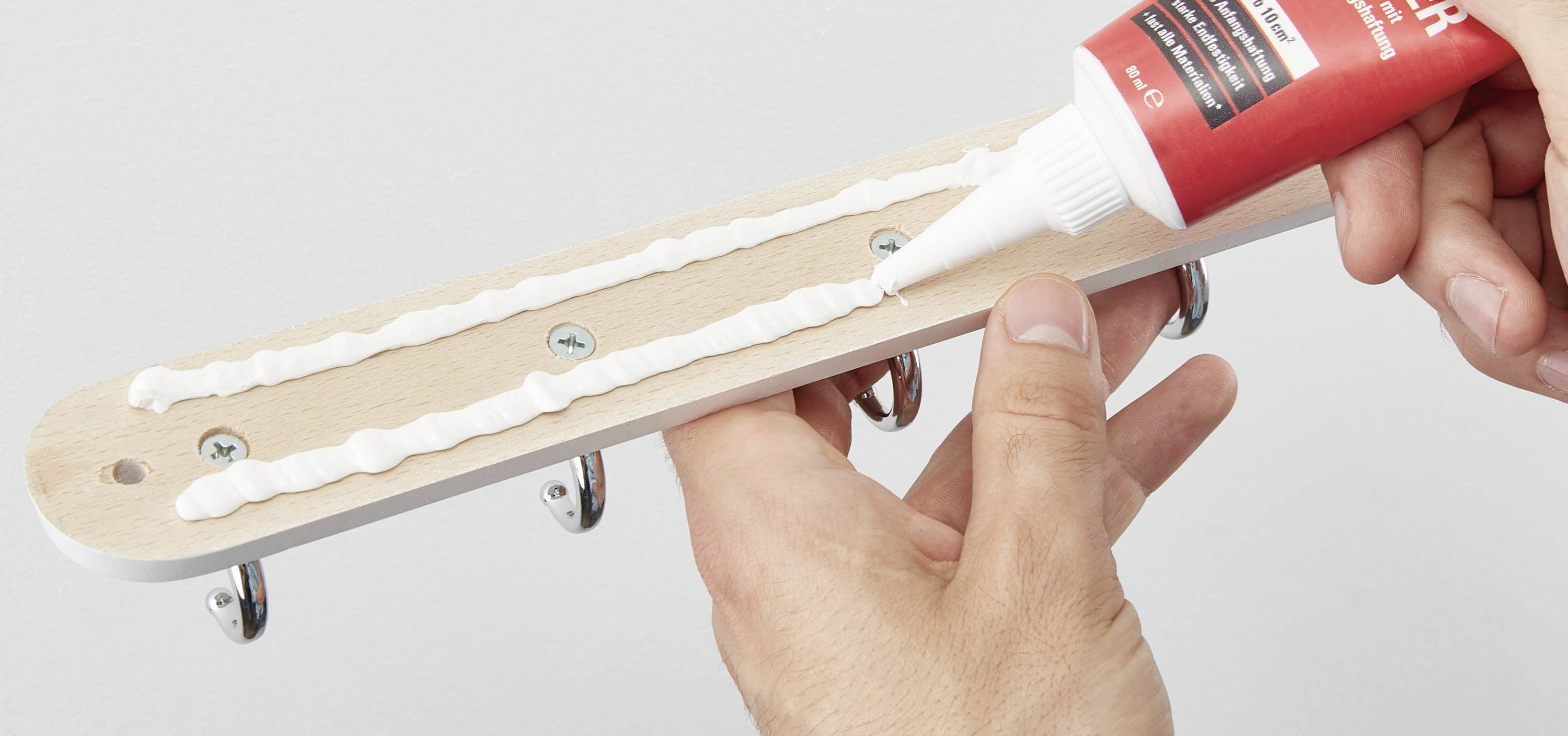 Eine Person trägt Klebstoff auf die Rückseite einer hölzernen Garderobenleiste mit Haken auf, um sie an der Wand zu befestigen.