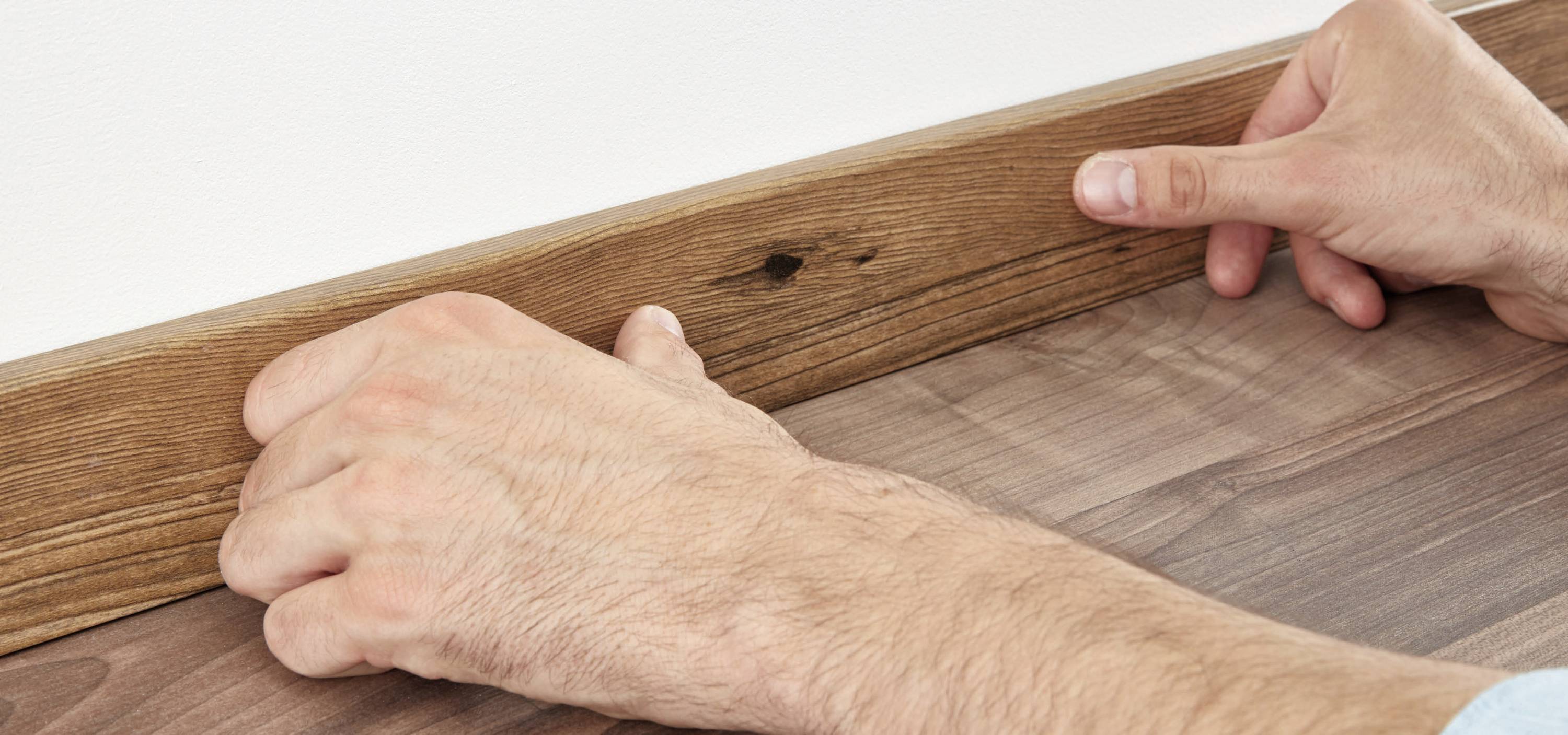 Zwei Hände montieren eine braune Holzleiste an einer Wand über einem Boden in ähnlichem Holzton.