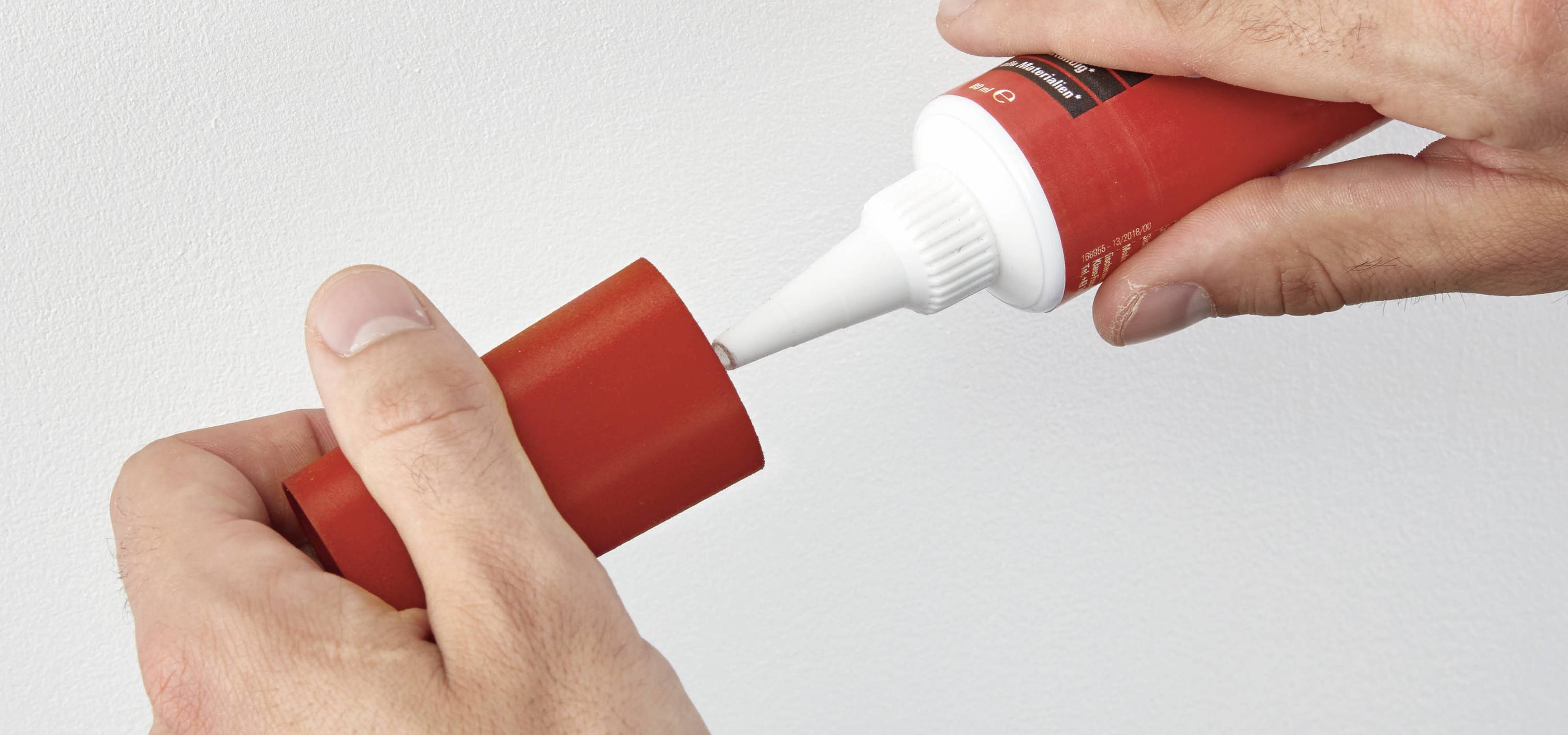 Zwei Hände öffnen eine rote Tube mit weißer Spitze. Möglicherweise handelt es sich um eine Dichtungs- oder Klebemasse.