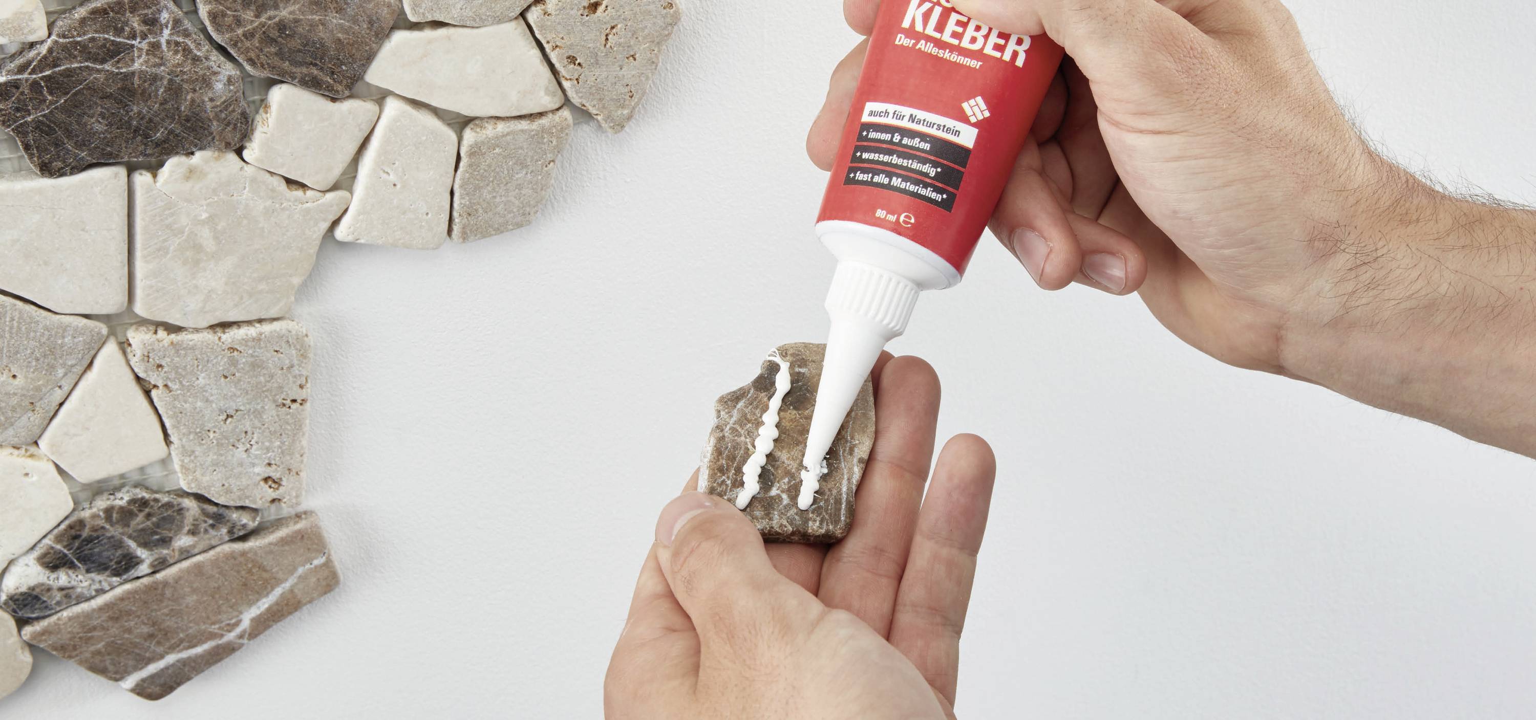 Eine Person trägt mit einer Tube einen weißen Kleber auf einen kleinen Stein auf, um Mosaikstücke an einer Wand zu befestigen.