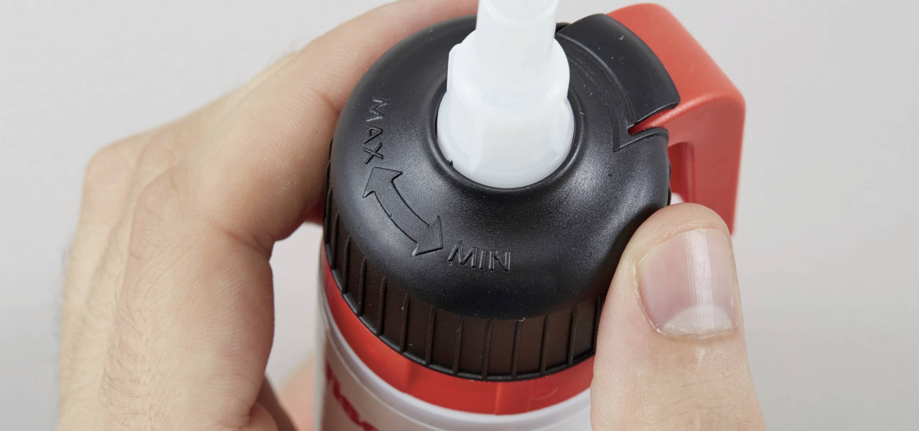 Eine Hand hält eine Spraydose, die an der Oberseite eine verstellbare Düse mit den Markierungen 'MAX' und 'MIN' zeigt.