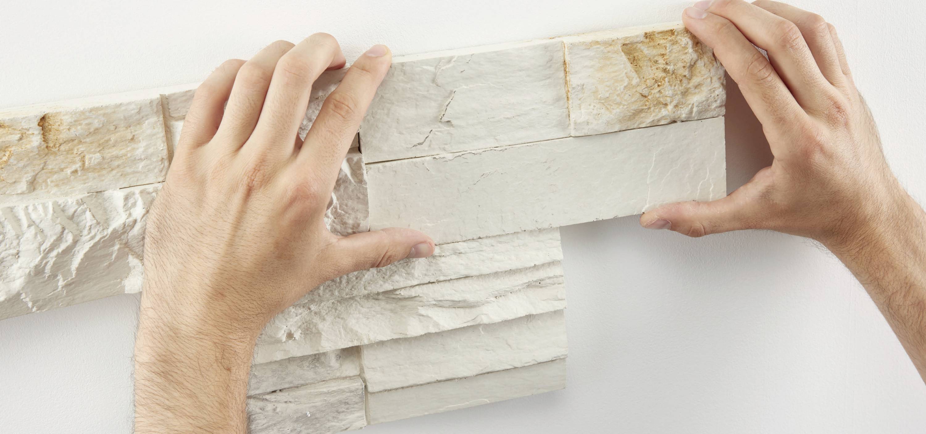 Zwei Hände verlegen weiße Wandfliesen in Steinoptik auf einer Wand.