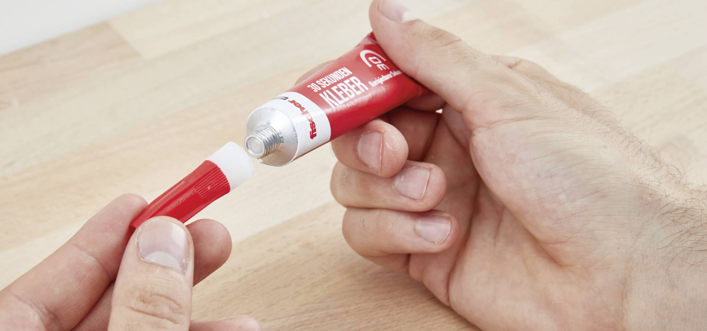 Eine Person öffnet eine rote Tube Wachsmalstift. Die Tube hat eine weiße Kappe und wird auf einem hölzernen Tisch gehalten.