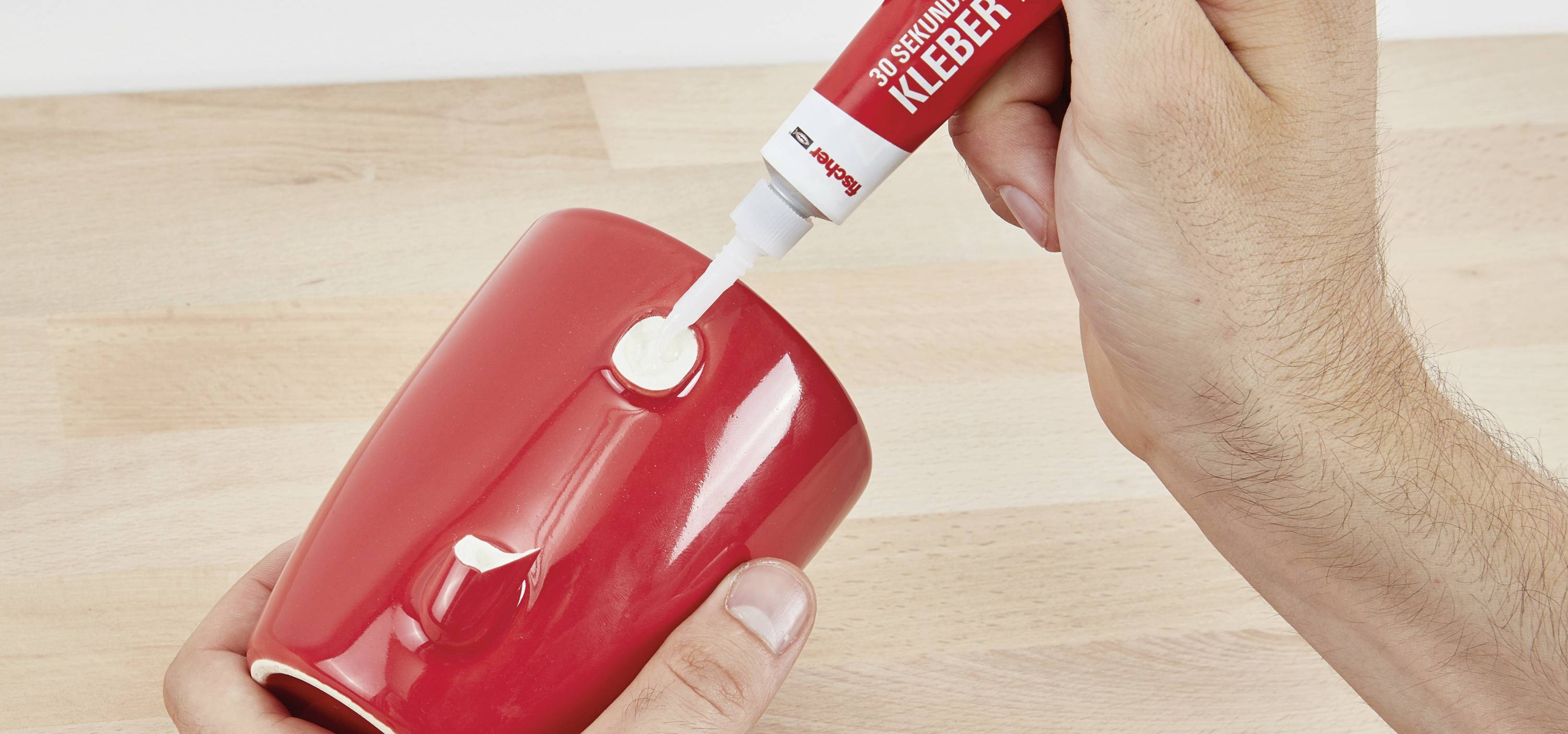 Eine rote Tasse wird mit einem weißen Kleber repariert. Eine Hand hält die Tasse, während die andere Kleber aus einer Tube aufträgt.