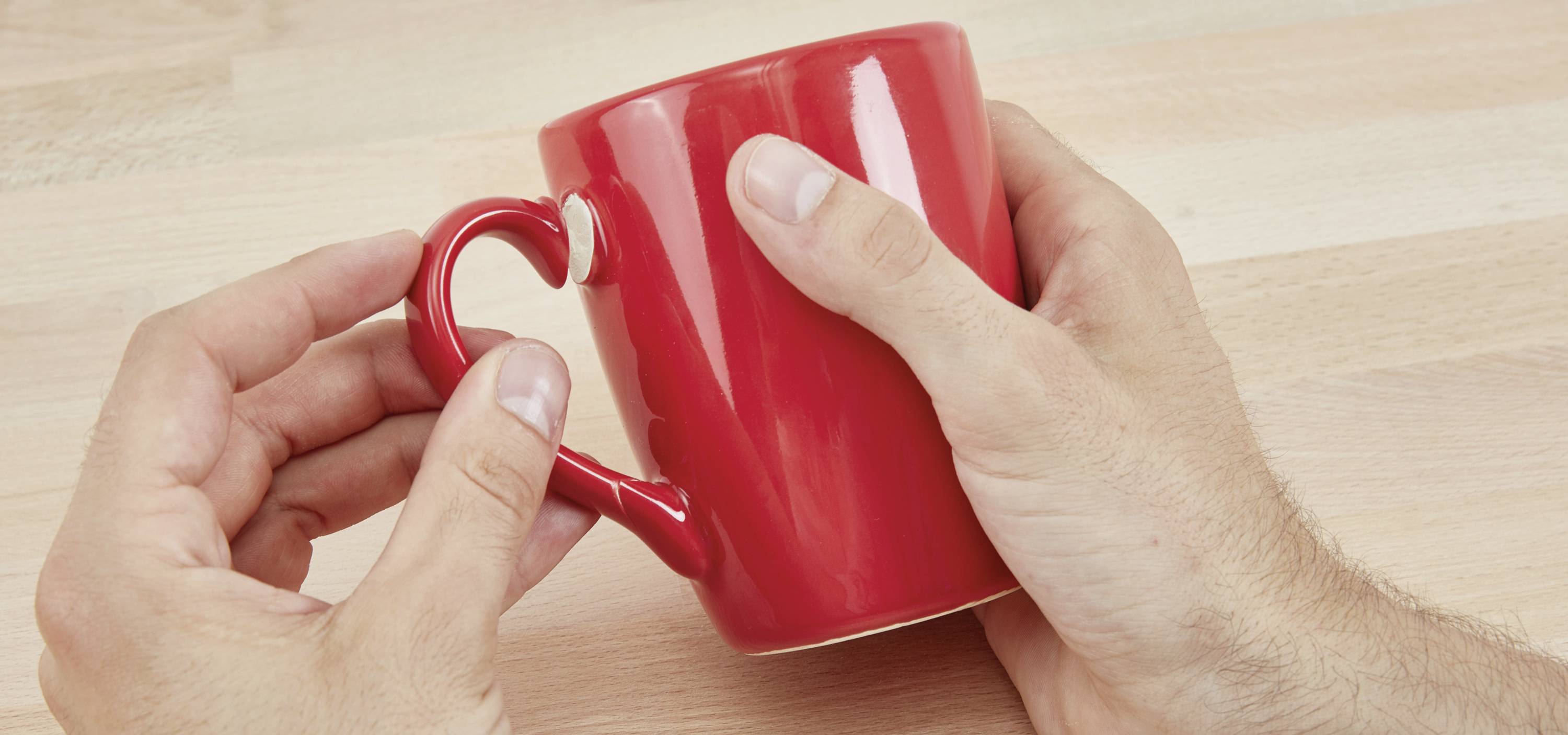 Zwei Hände halten eine rote Tasse mit herzförmigem Griff auf einem hölzernen Tisch.