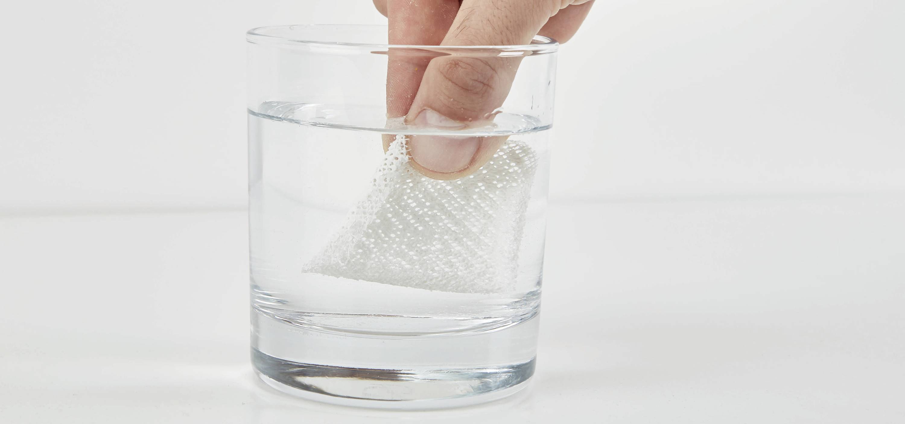 Eine Hand taucht einen weißen Reinigungsschwamm in ein Glas mit klarem Wasser.