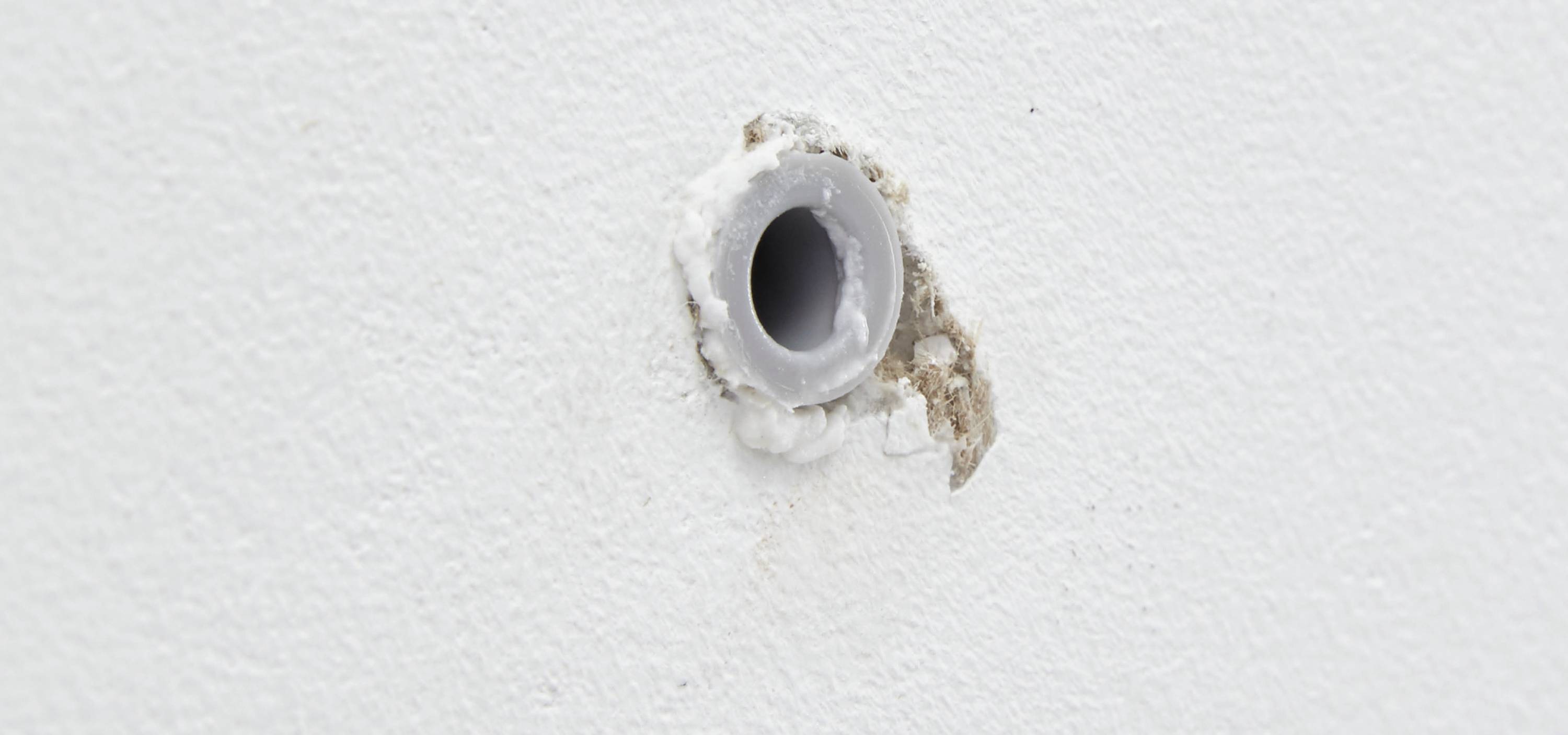 Nahaufnahme eines Lochs in einer weißen Wand, umgeben von Abnutzungsspuren. Möglicherweise für eine Halterung oder ein Kabel.