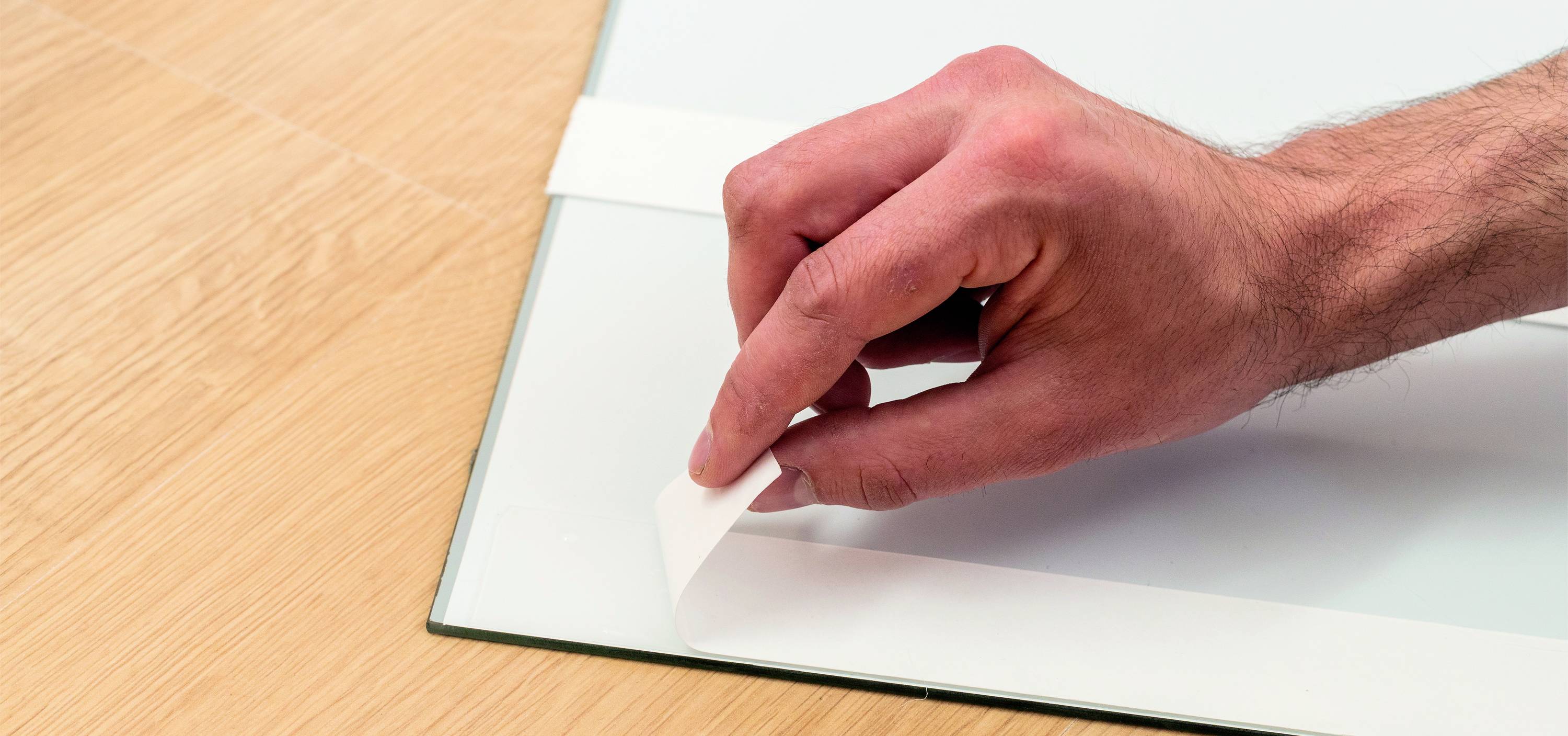 Eine Hand zieht einen Klebestreifen von einer Glasfläche ab, auf einem Holzboden liegend.