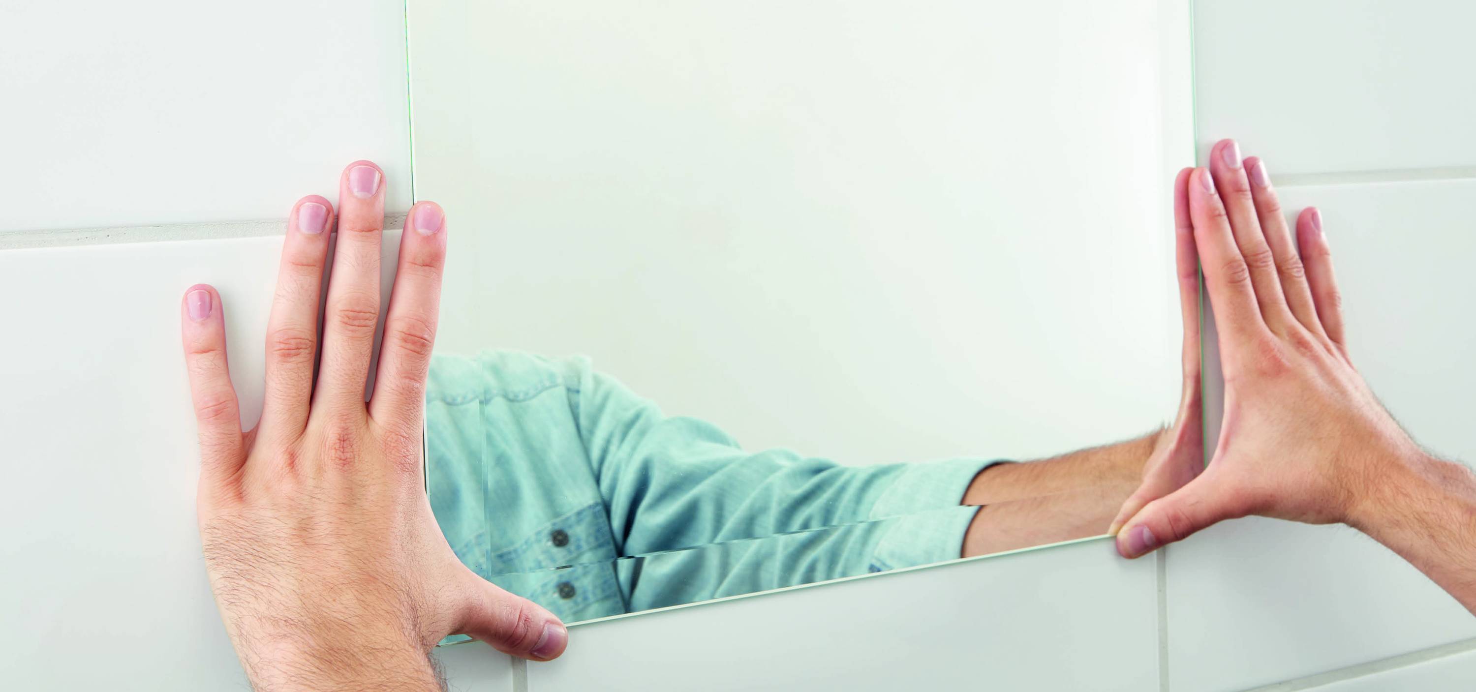 Eine Person richtet einen Spiegel an einer gefliesten Wand aus. Nur Hände und ein Teil des Oberkörpers sind sichtbar.