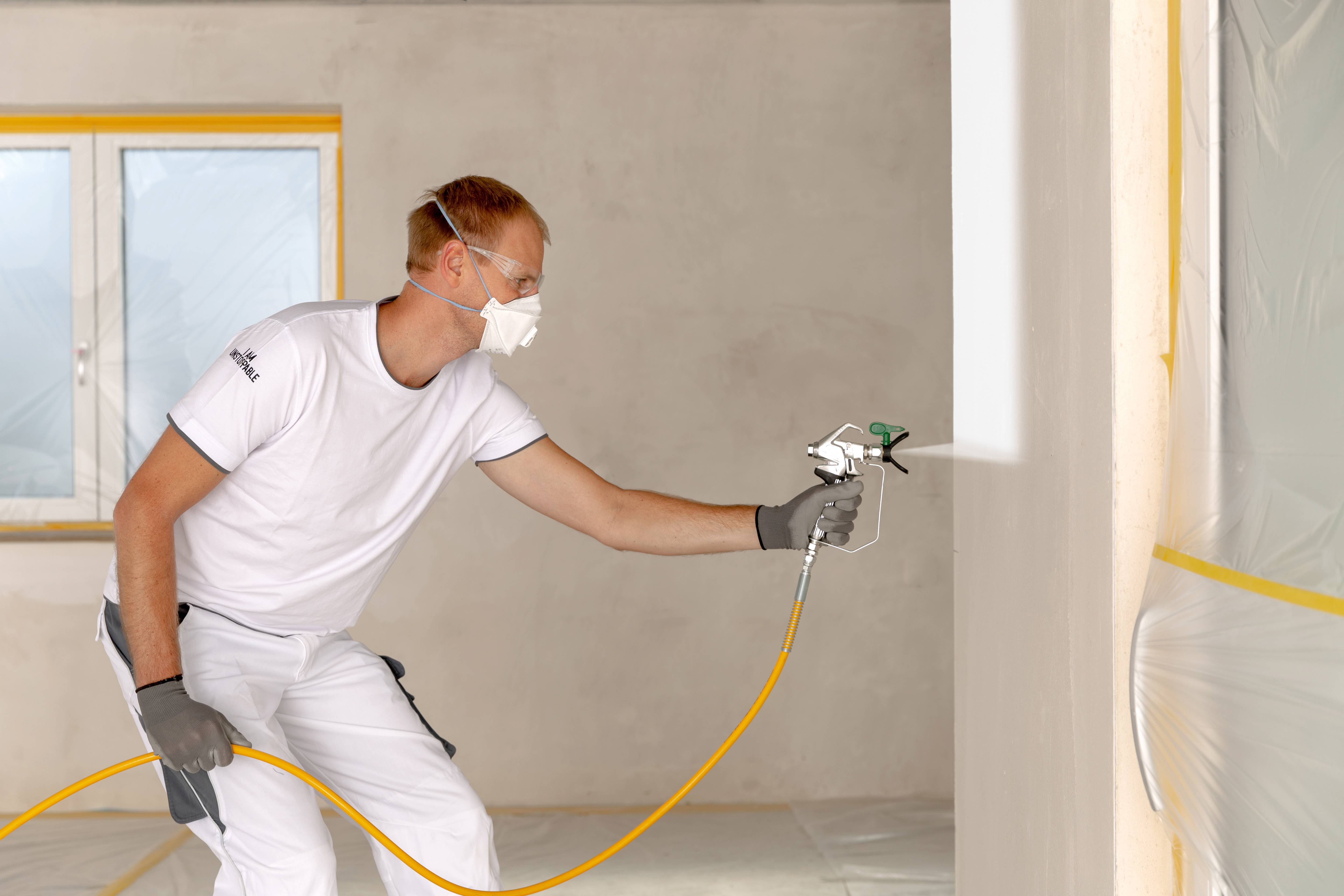 Ein Mann sprüht mit einer Spritzpistole Farbe an eine Wand in einem Raum. Er trägt Schutzkleidung und eine Maske.