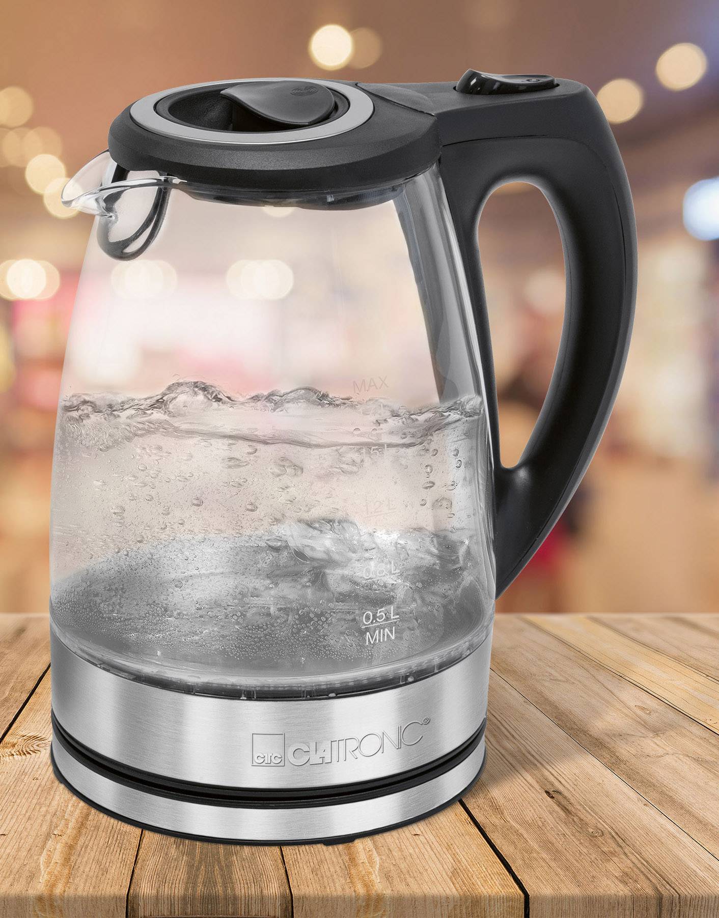 Wasserkocher mit schwarzem Griff und Glasgehäuse steht auf einem Holztisch. Wasser im Kessel kocht. Hintergrund ist unscharf.