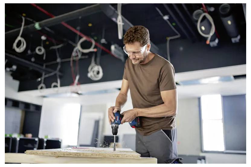 Ein Mann mit Sicherheitsschutzbrille bedient eine Bohrmaschine an einer Holzplatte auf einer Baustelle, wobei freiliegende Kabel von der Decke herunterhängen.