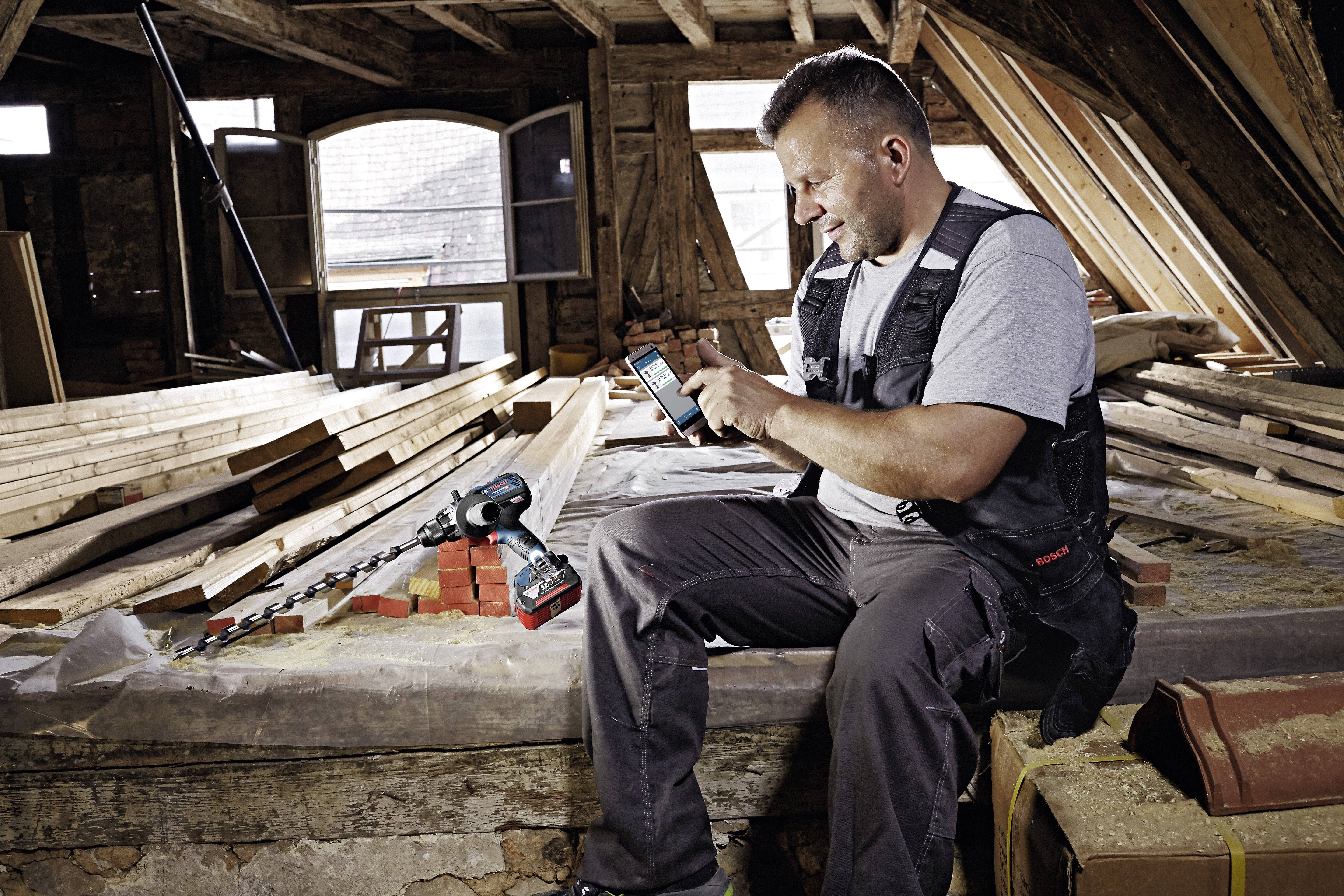 Ein Handwerker sitzt in einem Dachboden mit Holzmaterialien und Ziegelsteinen und überprüft etwas auf seinem Smartphone.