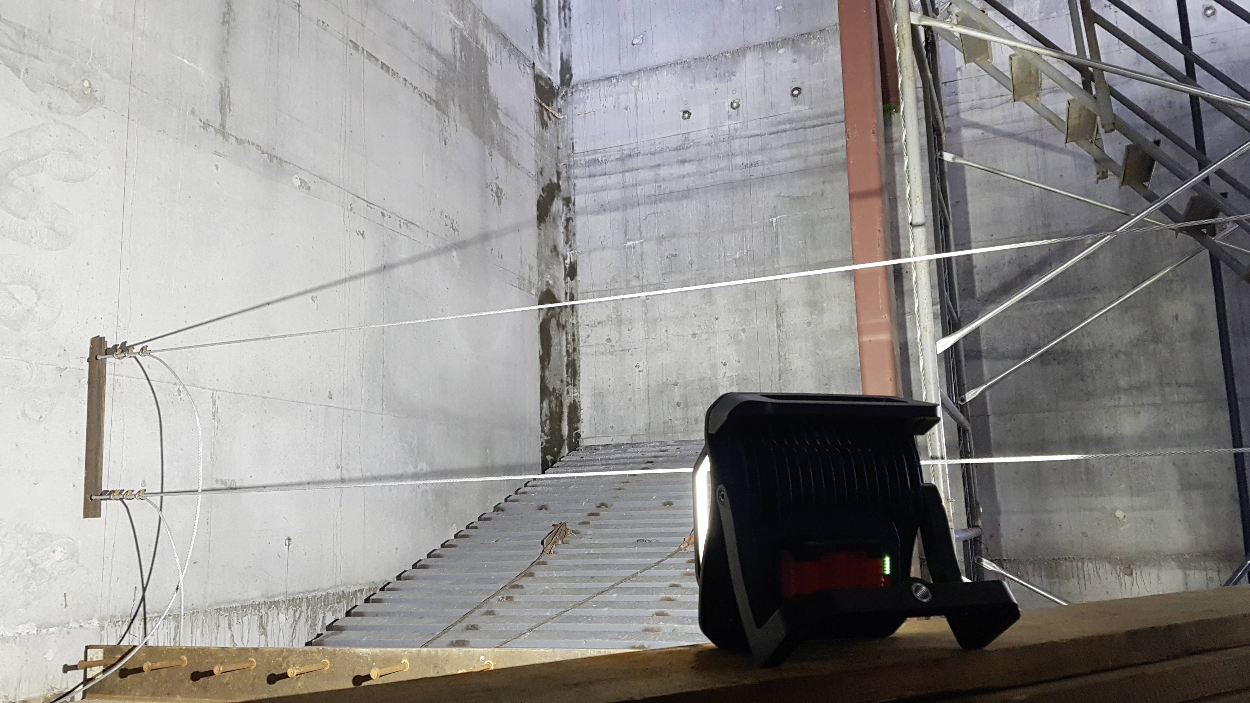 Ein Laser-Entfernungsmesser zielt auf eine große Betonwand in einem industriellen Raum. Metalltreppe und Stützen sind sichtbar.