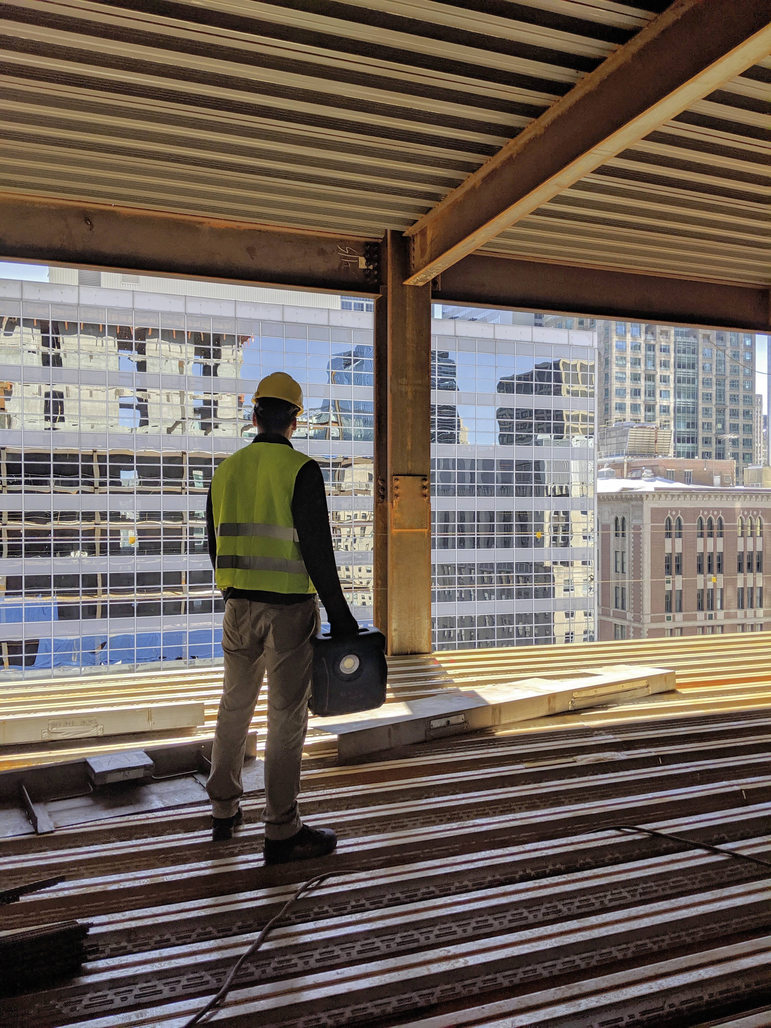 Ein Arbeiter mit Schutzhelm und Warnweste steht auf einer Baustelle und blickt auf eine Stadtsilhouette mit hohen Gebäuden.