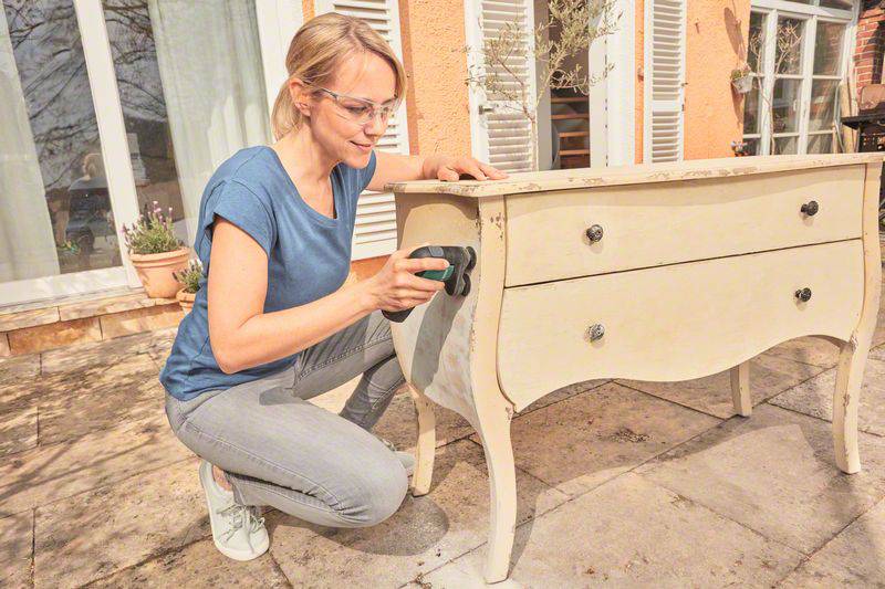 Eine Person schleift einen antiken Schrank auf der Terrasse, umgeben von Pflanzen und Fenstern.