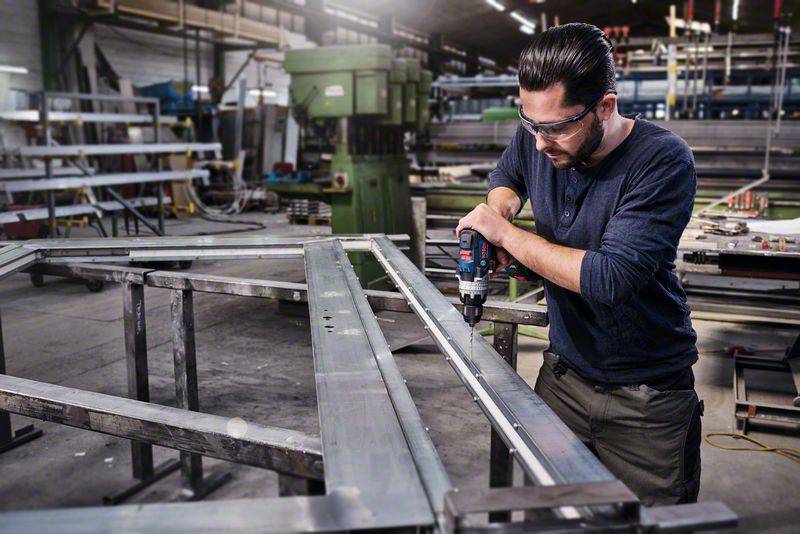 Ein Mann arbeitet in einer Fabrikhalle und bohrt mit einer elektrischen Bohrmaschine in ein Metallgestell.