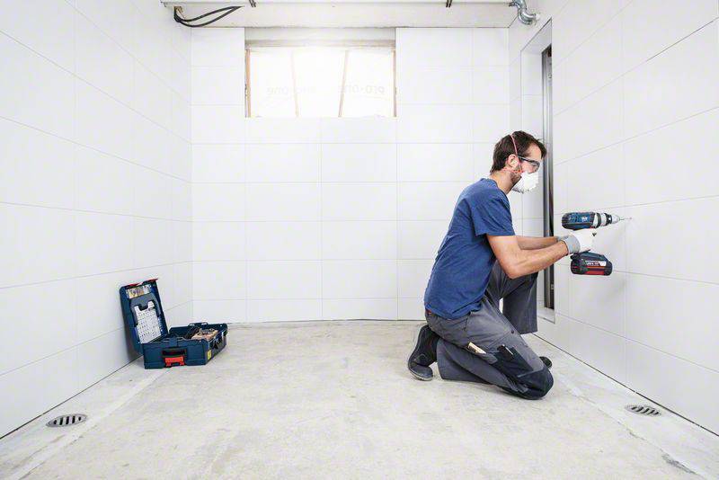 Ein Mann kniet auf dem Boden eines leeren Raums und verwendet eine Bohrmaschine an der Wand. Neben ihm liegt ein offener Werkzeugkasten.