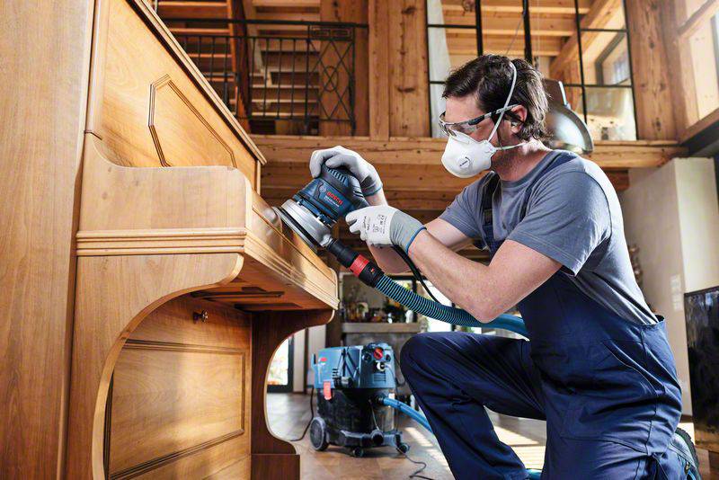 Ein Mann in Schutzkleidung schleift mit einer elektrischen Schleifmaschine die Oberfläche eines Holzpianos in einem holzgetäfelten Raum.