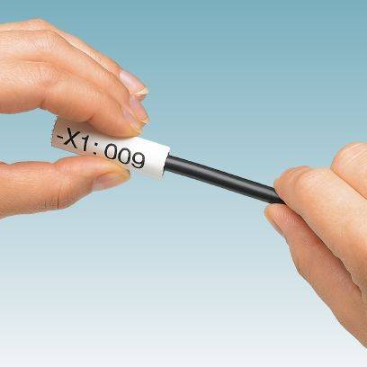 Zwei Hände halten ein schwarzes Kabel mit einem weißen Etikett, auf dem '-X1:009' steht. Der Hintergrund ist blau.