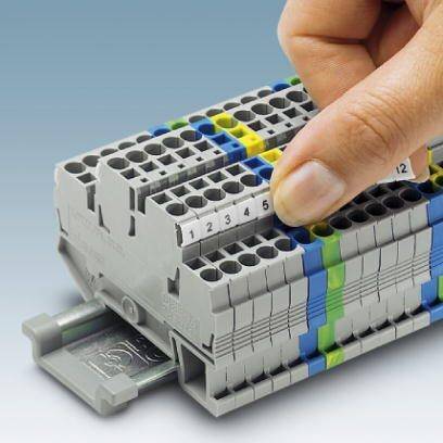 Eine Hand steckt einen nummerierten Stecker in eine graue, mehrfarbig markierte elektrische Klemmleiste.