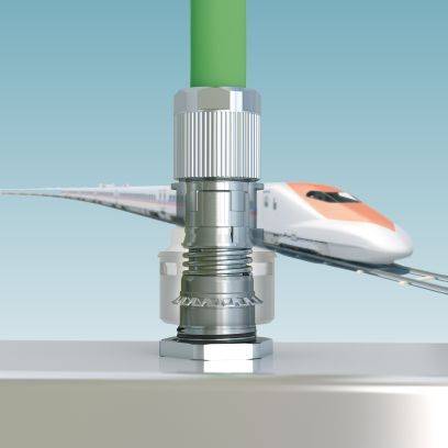 Ein Schnellzug rast im Hintergrund; im Vordergrund ein silberner Kompressionsadapter an einem grünen Rohr auf grauer Oberfläche.