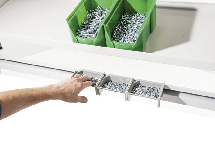 Eine Hand greift nach kleinen Metallteilen aus einem grauen Behälter unterhalb zweier grüner Kisten mit weiteren Schrauben.