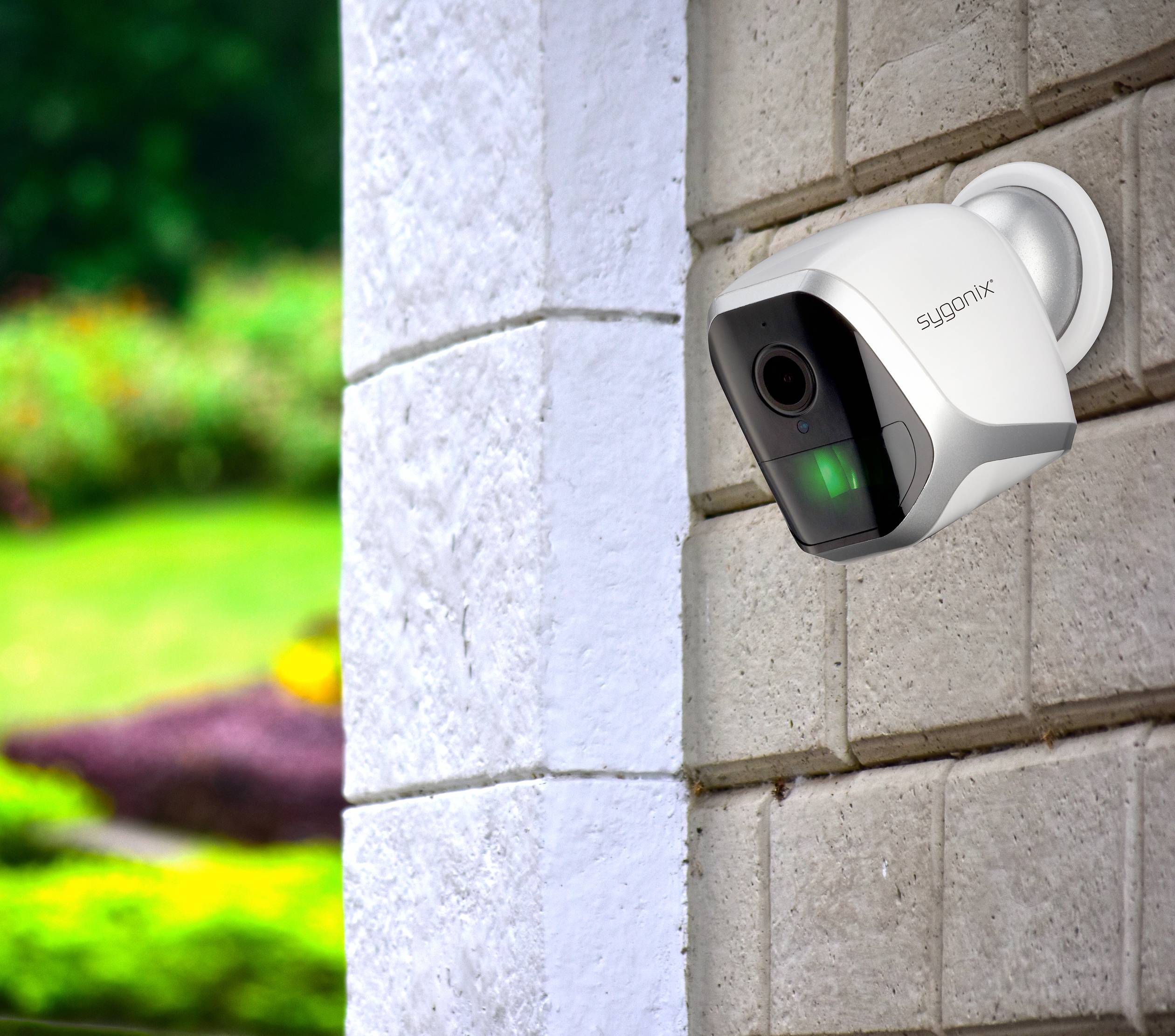Eine Überwachungskamera ist an einer Ziegelwand montiert, überwacht einen grünen Garten im Hintergrund.