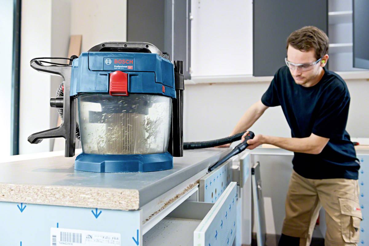 Ein Mann reinigt mit einem Industriesauger eine Küchenarbeitsplatte. Der Sauger ist blau und steht auf der Arbeitsfläche. Der Mann trägt Schutzbrille.