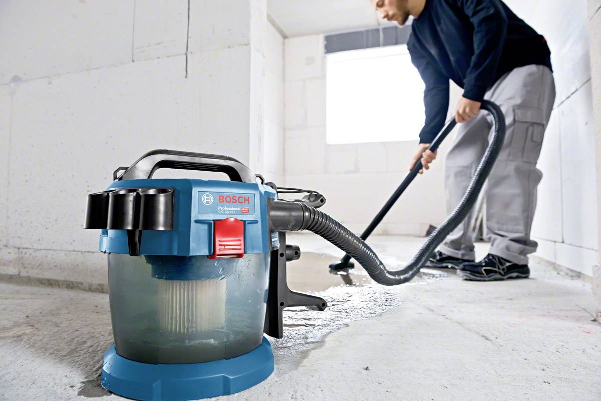 Ein Mann reinigt mit einem Bosch Bau-Staubsauger einen Betonboden in einem hellen Raum. Der Staubsauger ist blau und transparent.