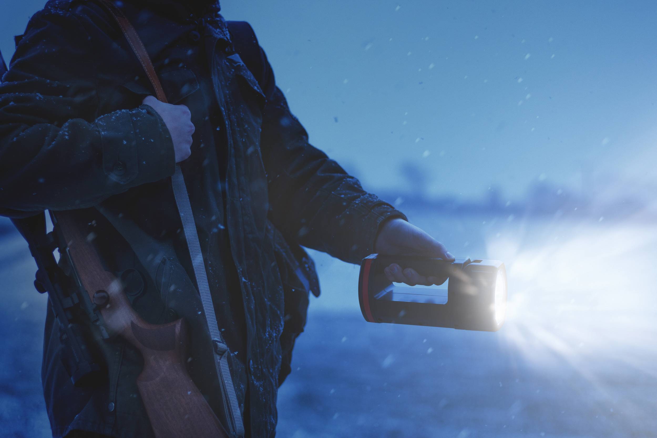Person mit Jagdgewehr und Taschenlampe im Schnee, erhellt die Umgebung bei Dämmerung.