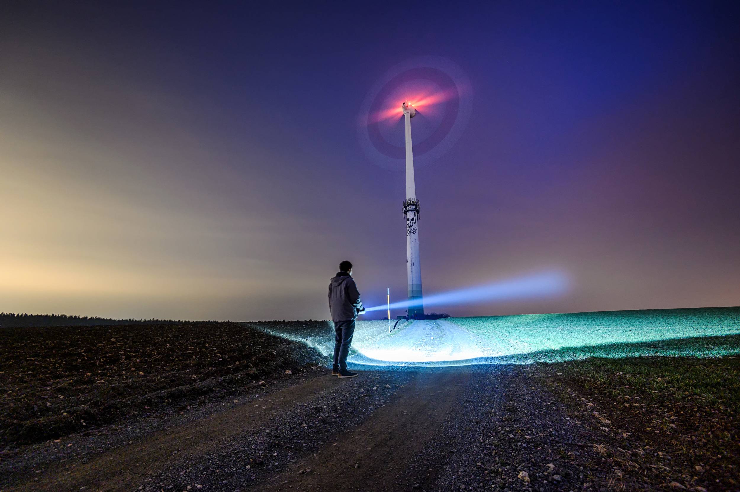 Eine Person steht bei Nacht auf einem Feldweg und betrachtet ein beleuchtetes Windrad mit roten und blauen Lichtern am Himmel.