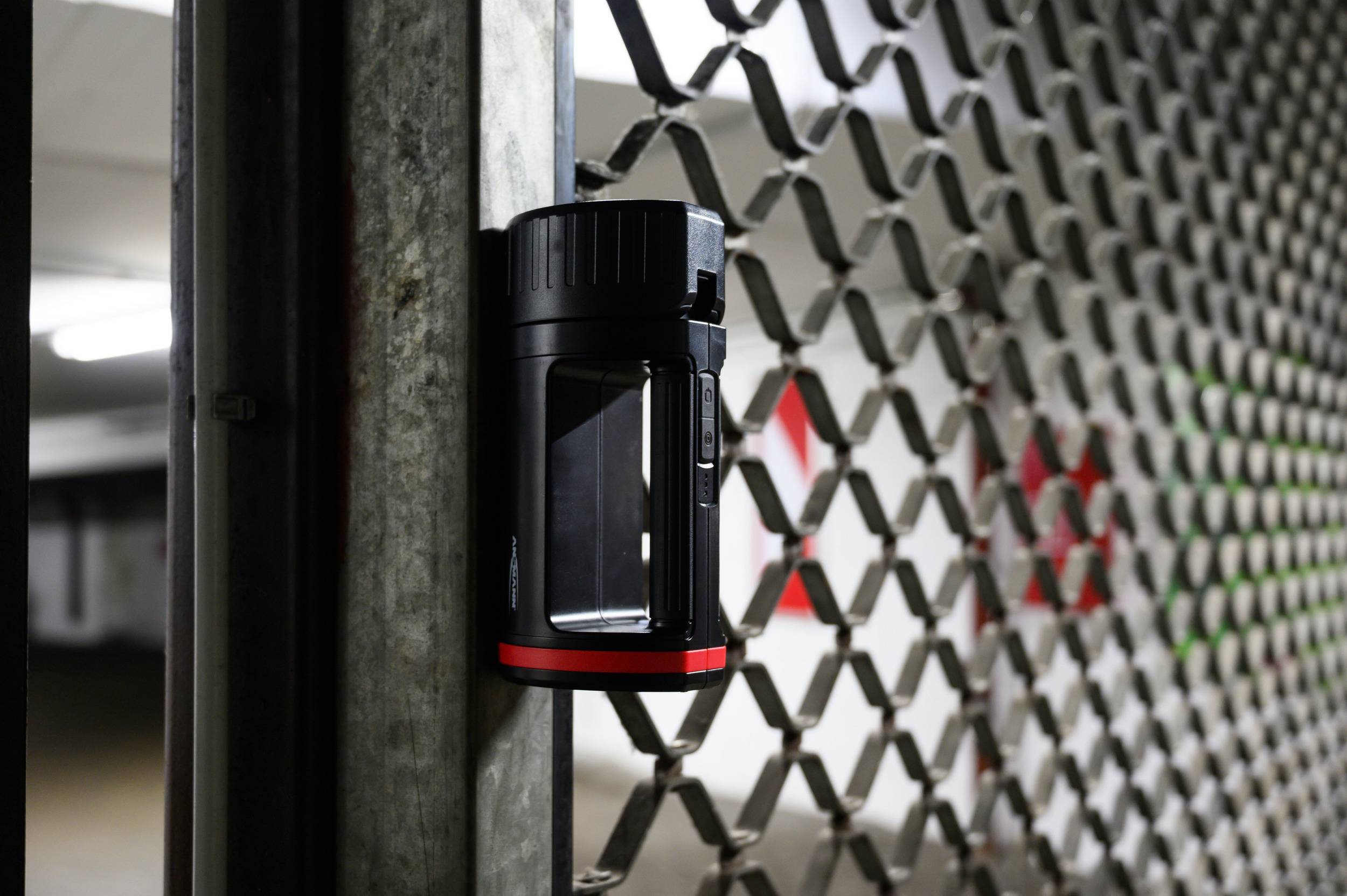 Eine schwarze und rote Magnettaschenlampe haftet an einem Gitter in einem Parkhaus. Im Hintergrund sind unscharfe Lichter zu sehen.