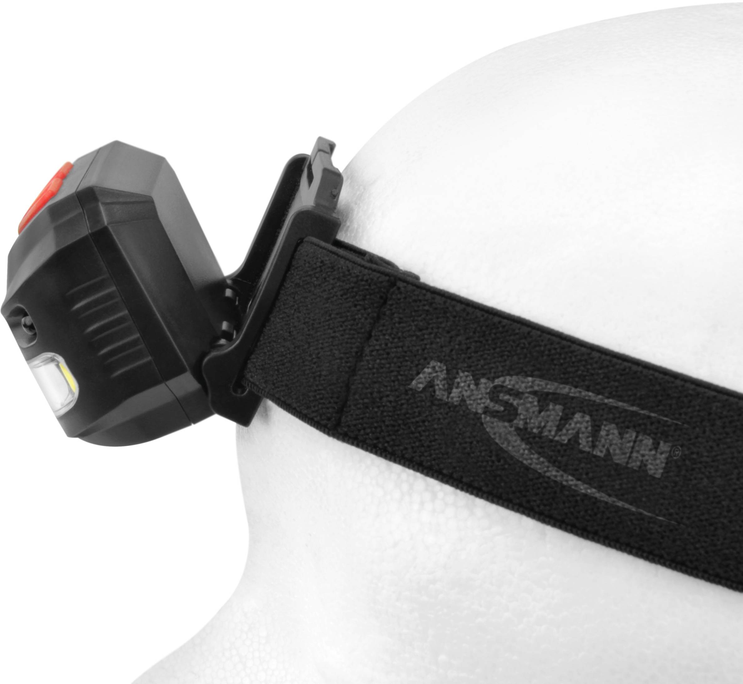 Stirnlampe auf Kopfmodell, schwarzes Band mit 'ANSMANN' Logo, rechteckige Lampe mit rotem Knopf und LED-Leuchte.