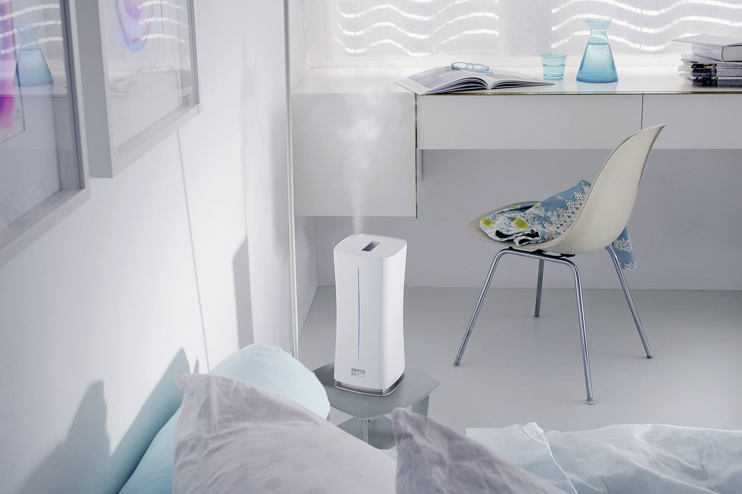 Luftbefeuchter in modernem, hellem Schlafzimmer, steht auf einem kleinen Tisch neben dem Bett. Auf einem Schreibtisch im Hintergrund liegen Bücher.