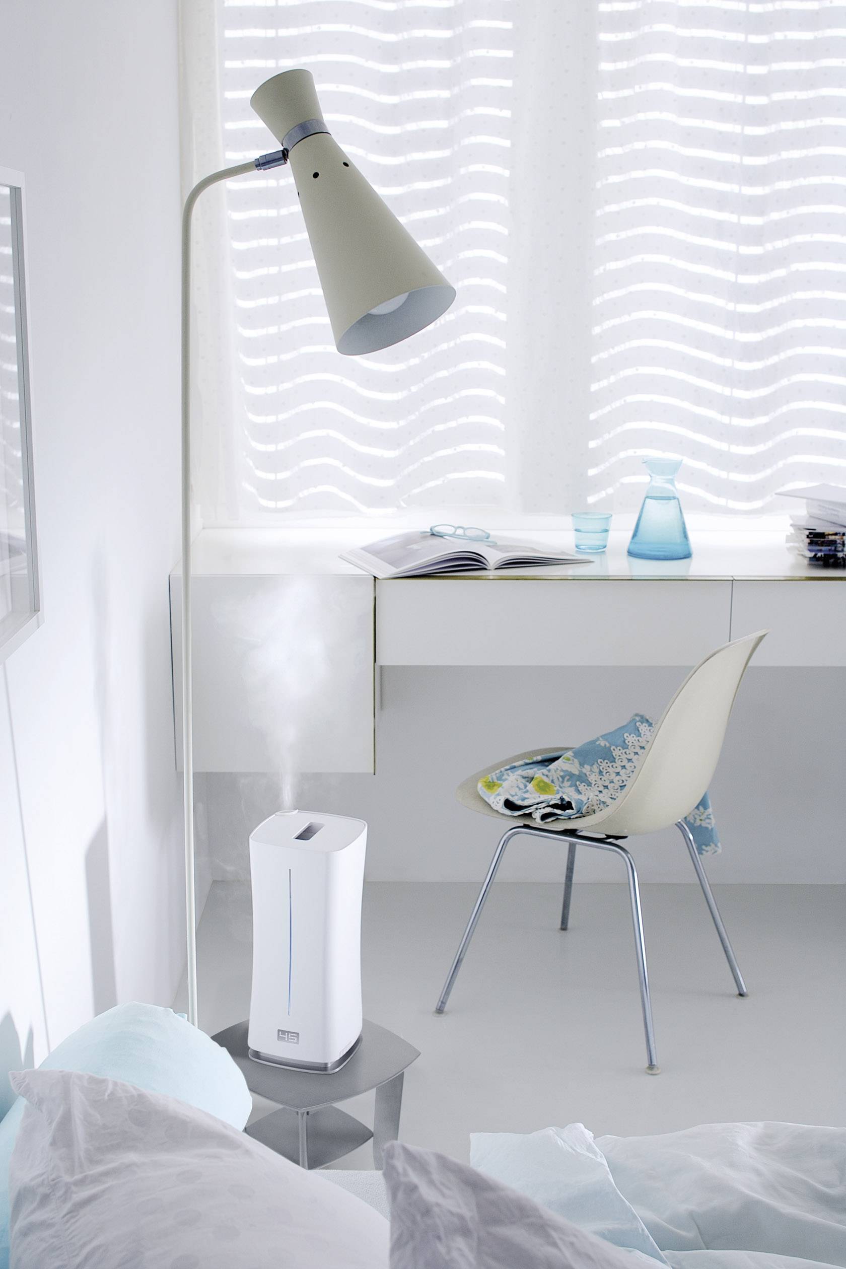 Ein moderner Raum mit weißem Schreibtisch, darauf liegen ein offenes Buch und ein blaues Glas. Ein Stuhl daneben, Lampe beleuchtet. Ein Luftbefeuchter steht links.
