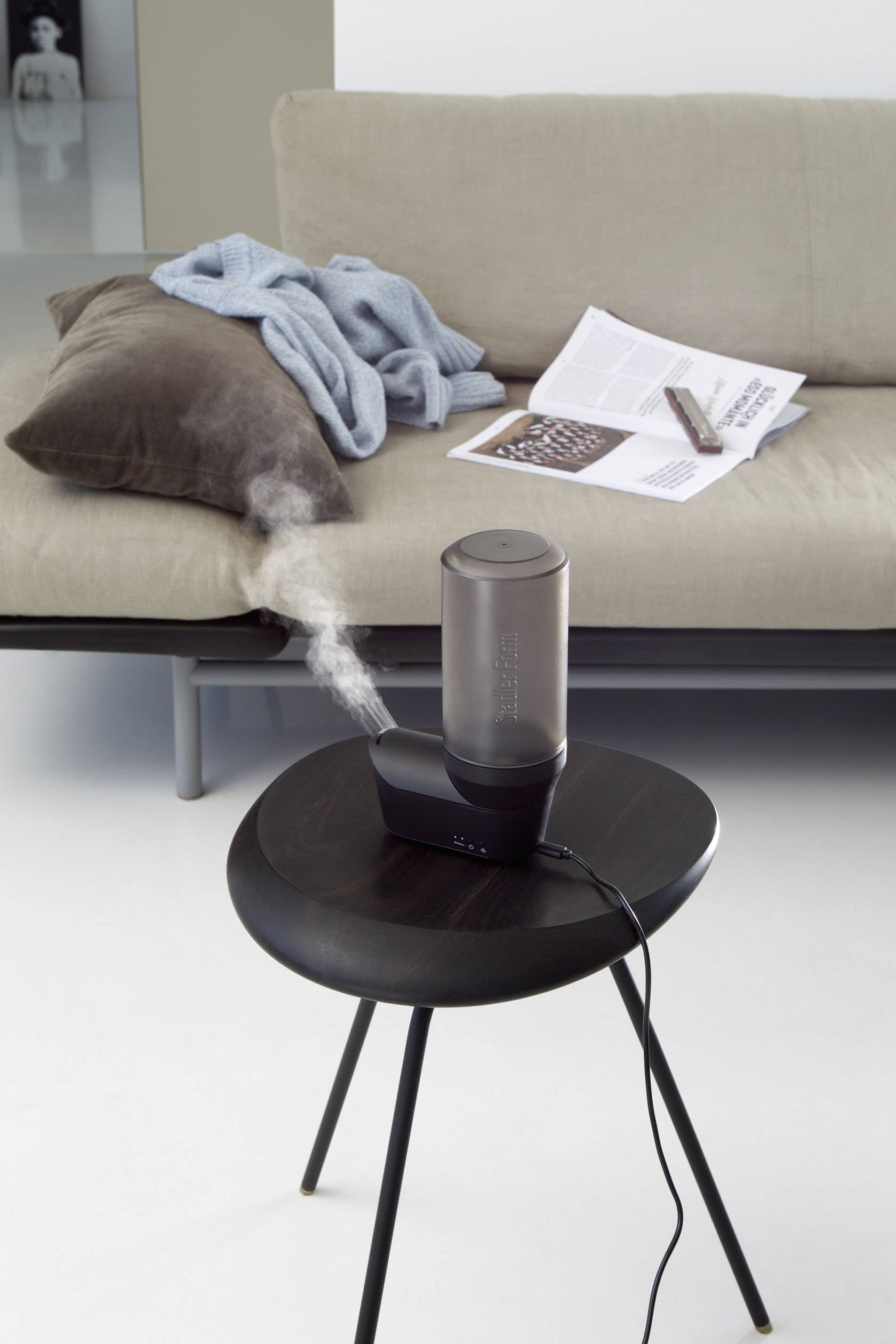 Ein Tischventilator steht auf einem kleinen Tisch, neben einem Sofa mit Decken und einer offenen Zeitung; der Raum wirkt modern und gemütlich.