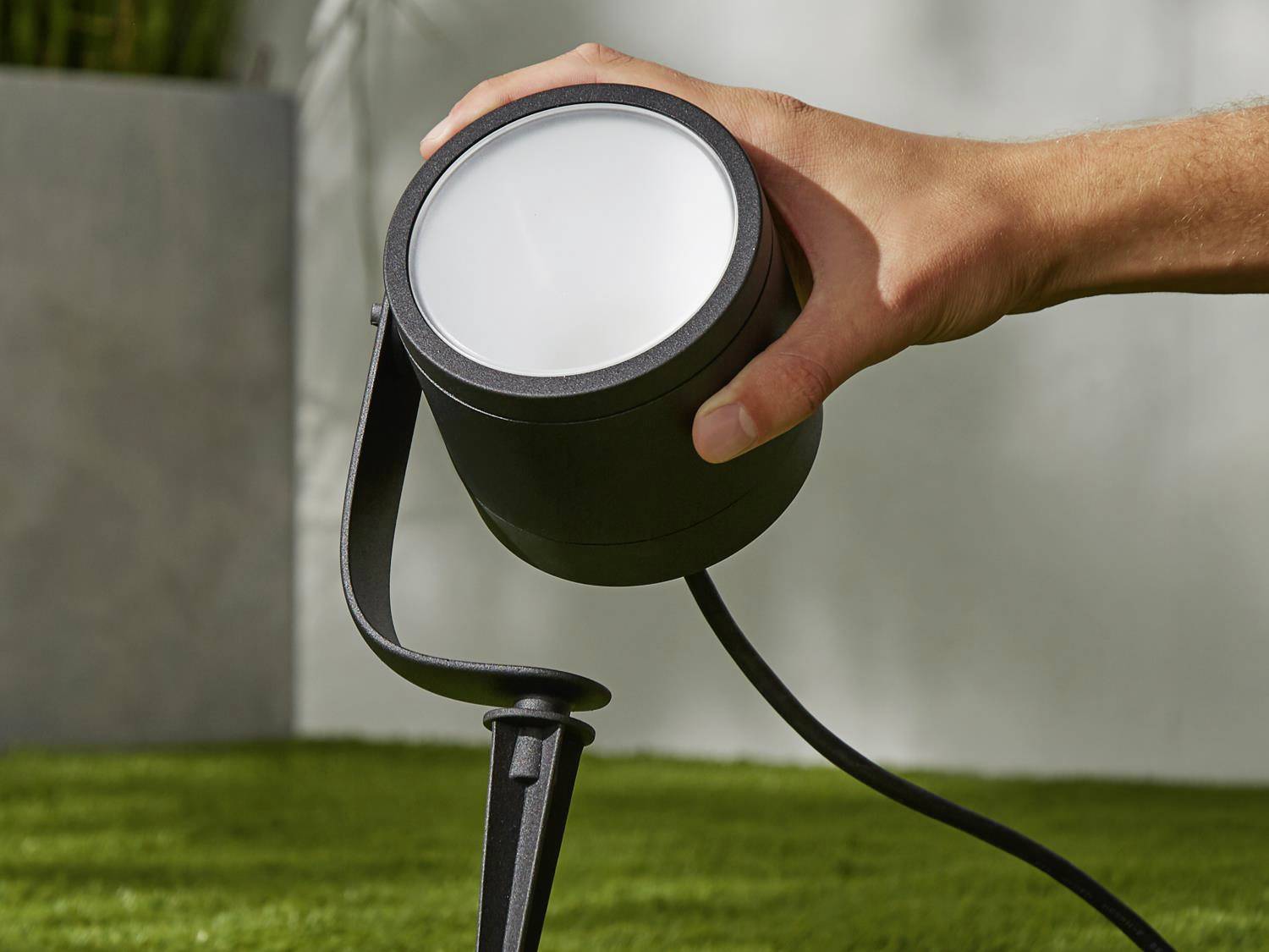 Eine Hand richtet ein schwarzes Außenstrahlerlicht auf grünem Gras aus. Der Fokus liegt auf dem Strahler und der Handbewegung.