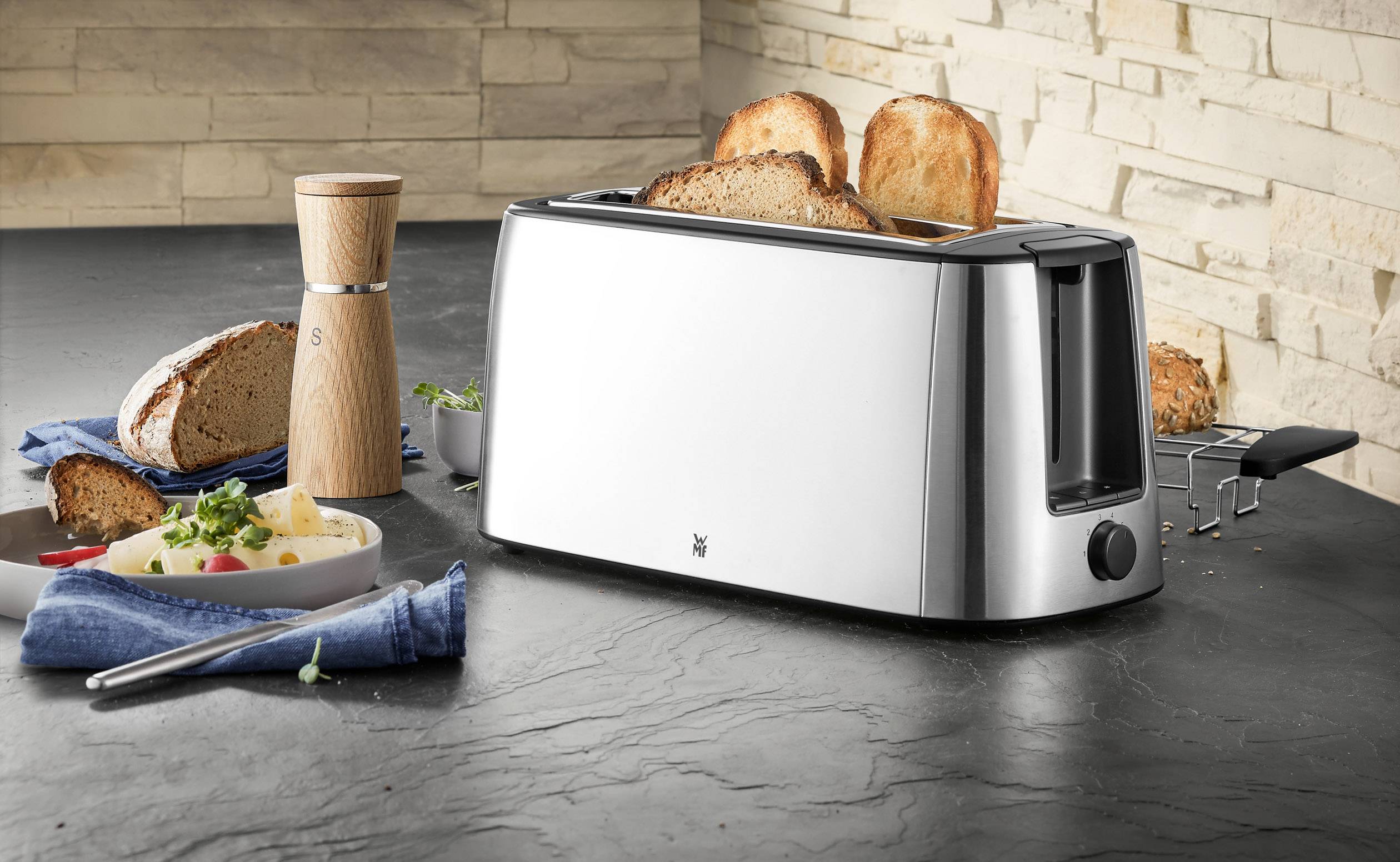 Silberner Toaster mit zwei getoasteten Brotscheiben auf einer Arbeitsplatte, daneben geschnittenes Brot, ein Holzpfeffermühle und ein Teller mit Essen.